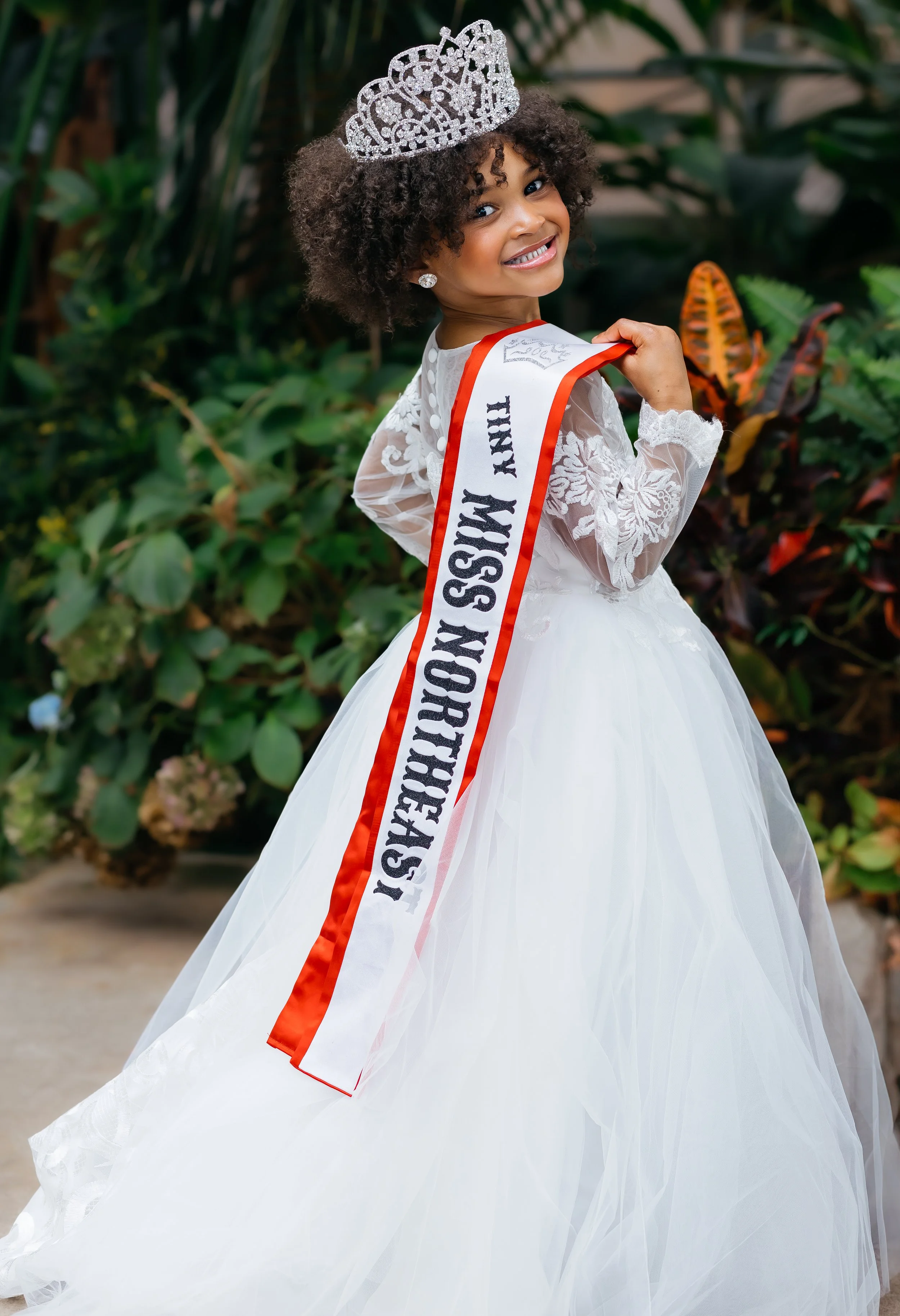 Young girl in a white lace dress with a crown and sash that reads "Miss Northfire XVI", smiling and looking back over her shoulder, standing outdoors with green foliage in the background.