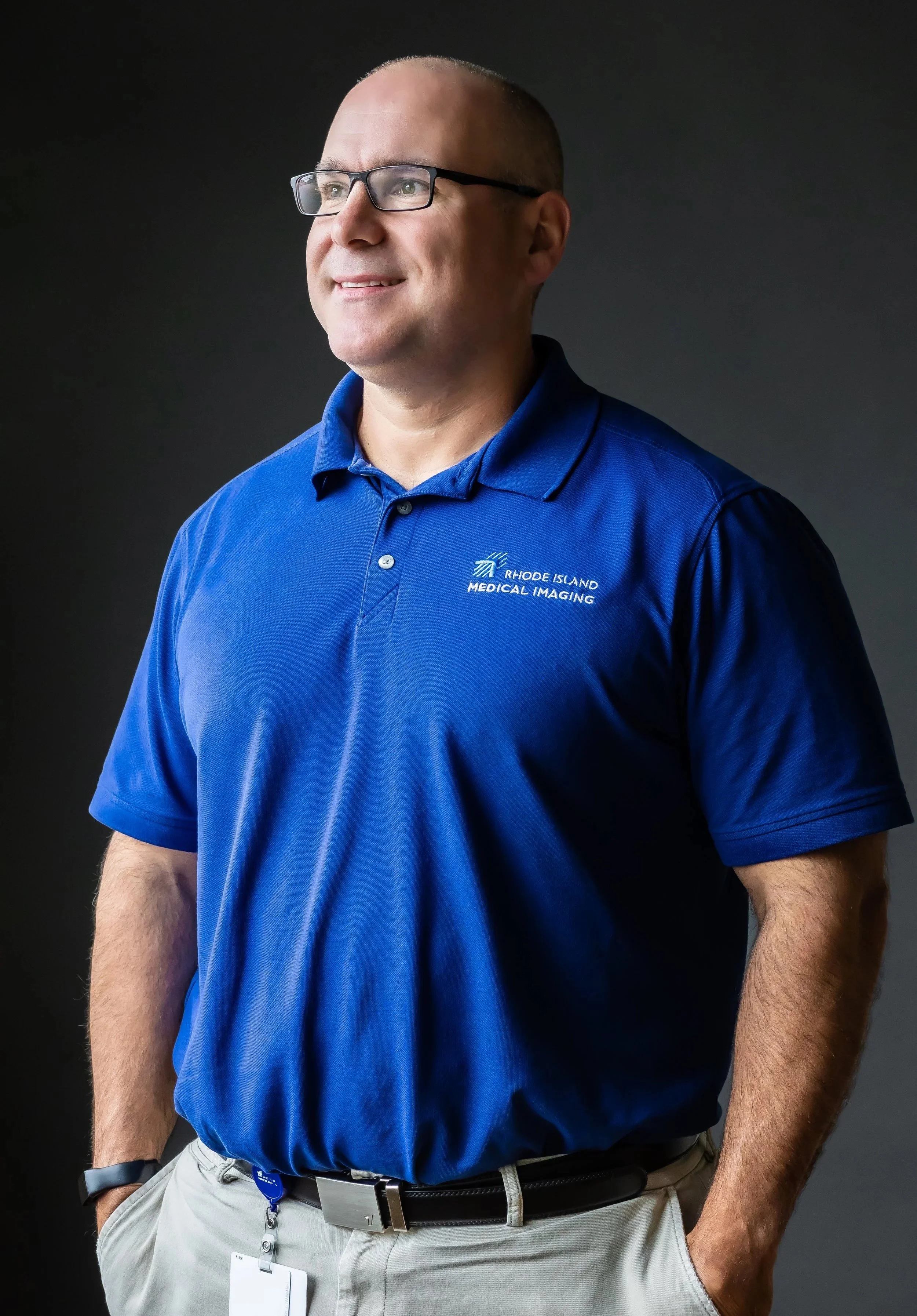 Man in blue polo shirt with 'Rhode Island Medical Imaging' logo, wearing glasses, standing against a plain dark background.