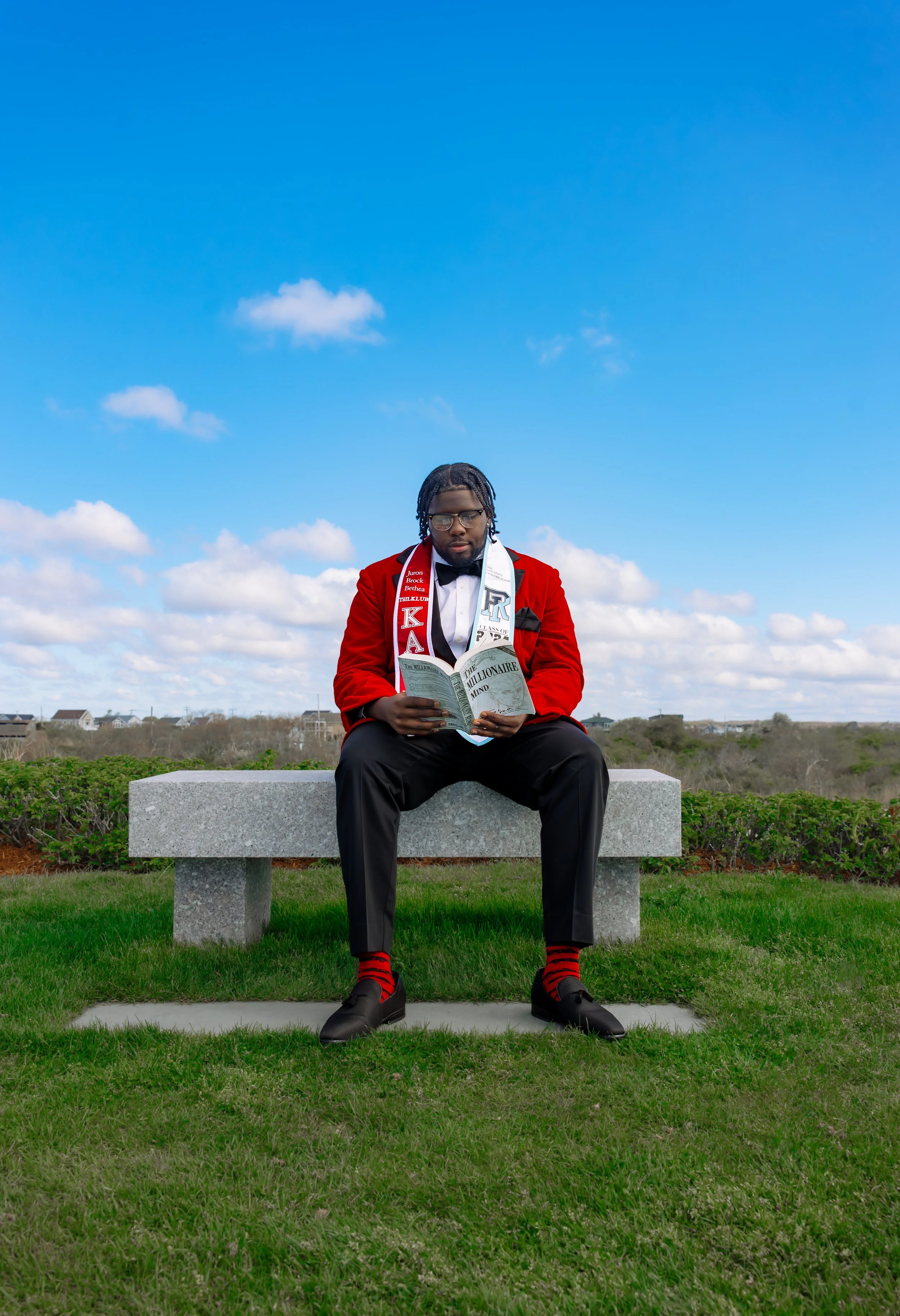 A man in a red blazer, black pants, and red and black socks sits on a stone bench outdoors, reading a magazine with a partly cloudy sky in the background.