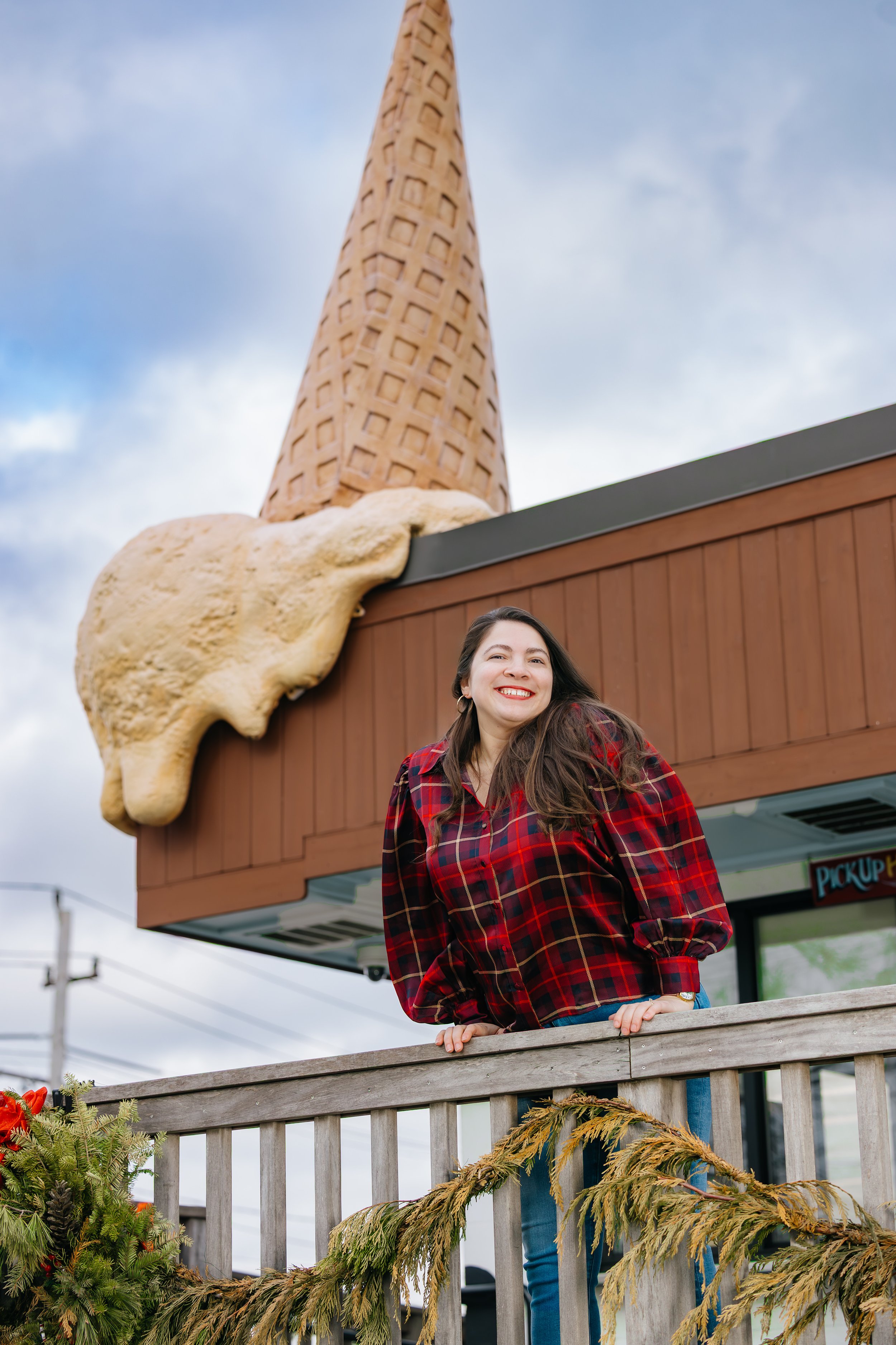 Woman in red plaid shirt leaning on a wooden railing with a large ice cream cone sculpture behind her at a themed location.