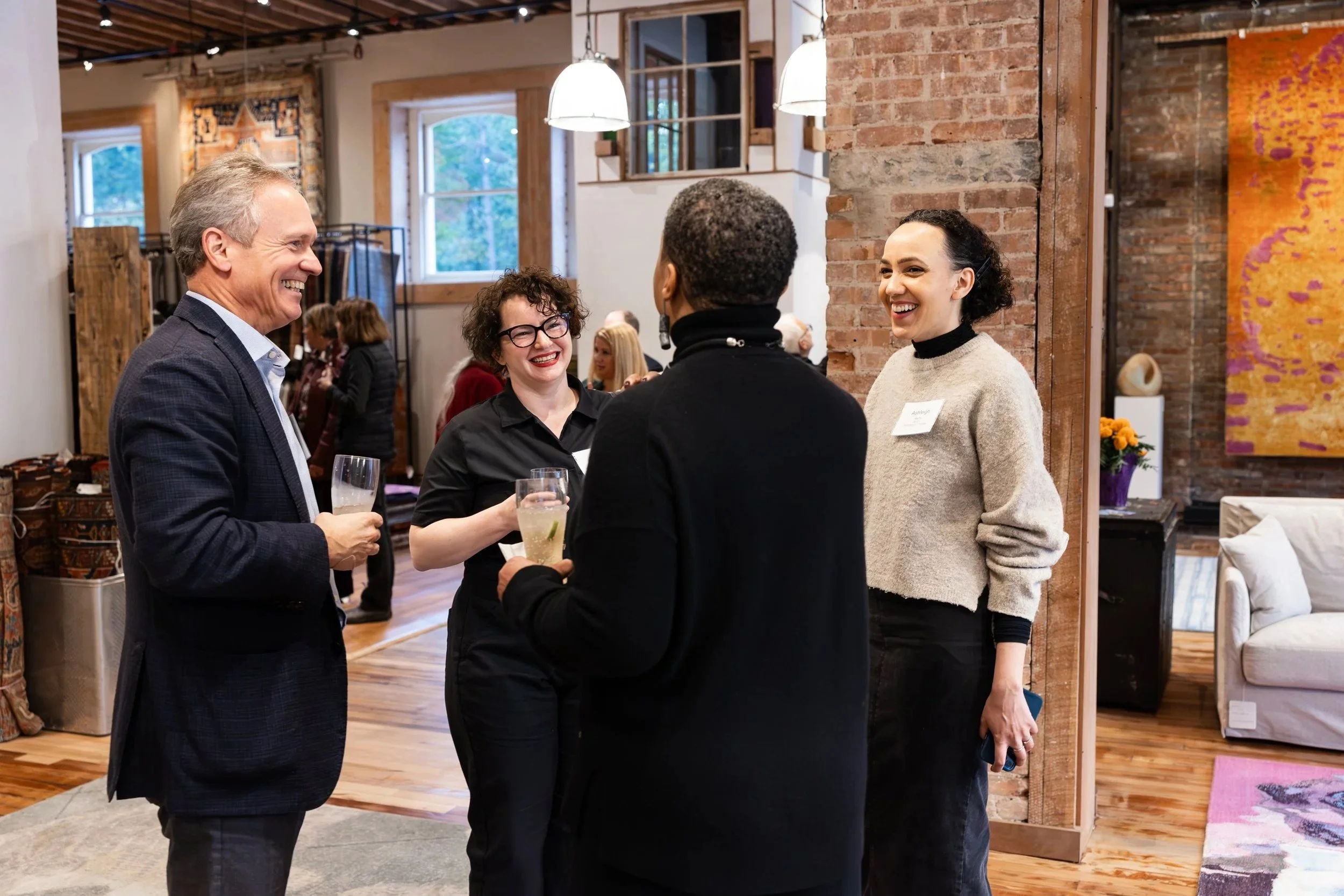 Four people are engaged in conversation and smiling at a social gathering in a cozy, art-filled indoor space with wooden floors and brick walls. Two of them are holding drinks.