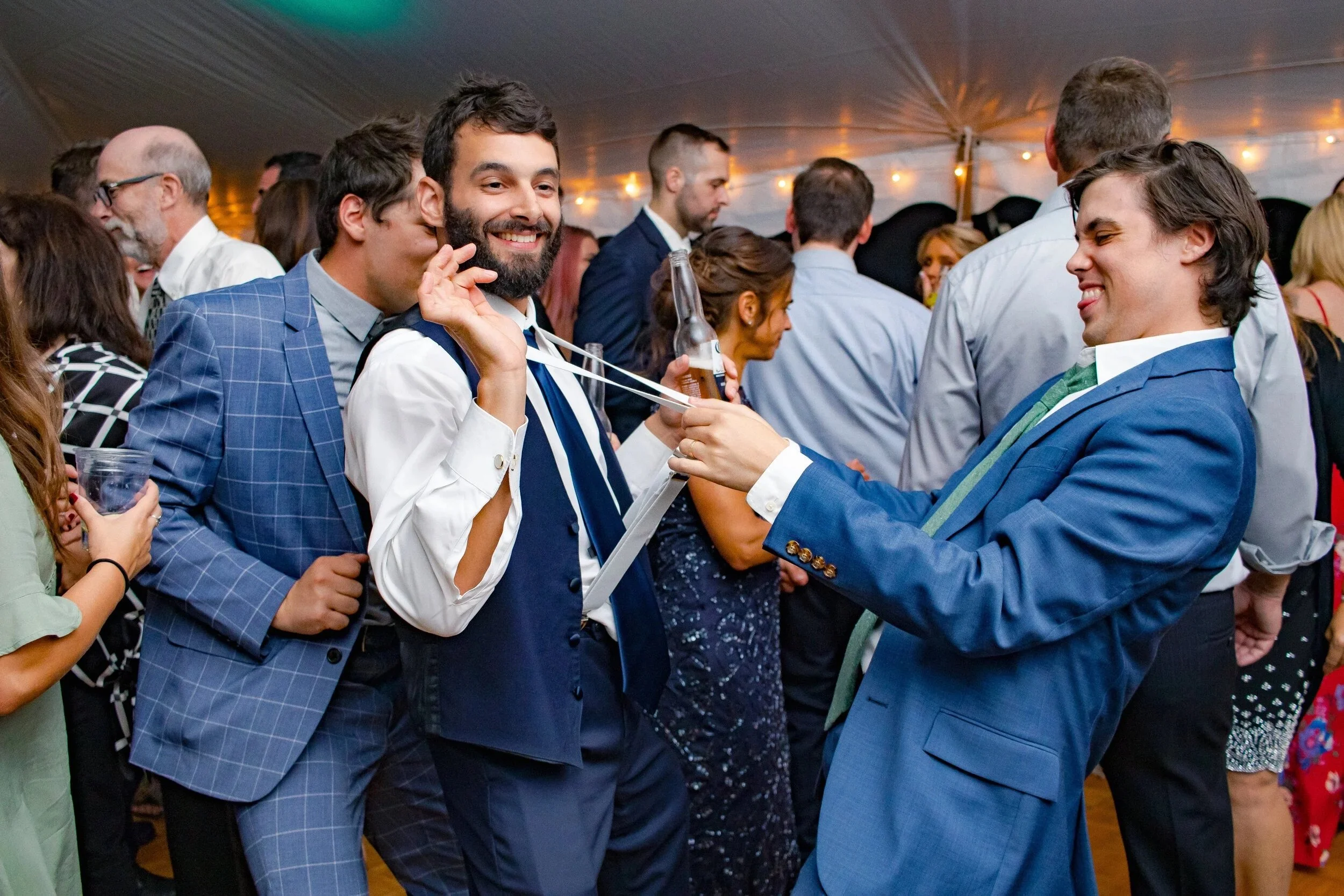 Group of people at a social gathering or party, with two men in the foreground playfully holding a drink and smiling, under a tent decorated with string lights.