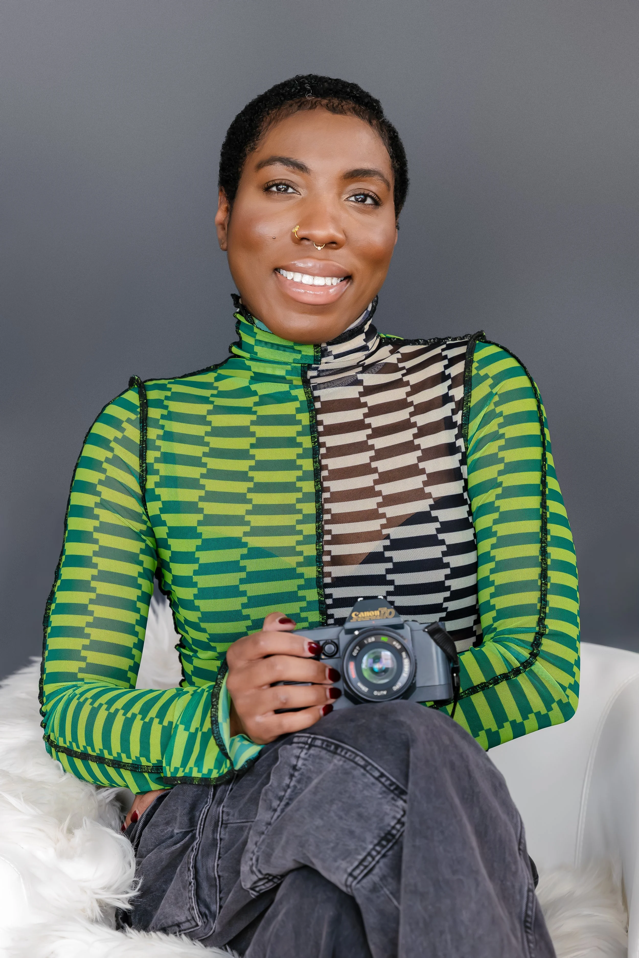 A smiling woman with short curly hair holding a camera. She is wearing a colorful patterned top with green, black, and white designs, and sitting on a white furry chair against a gray background.