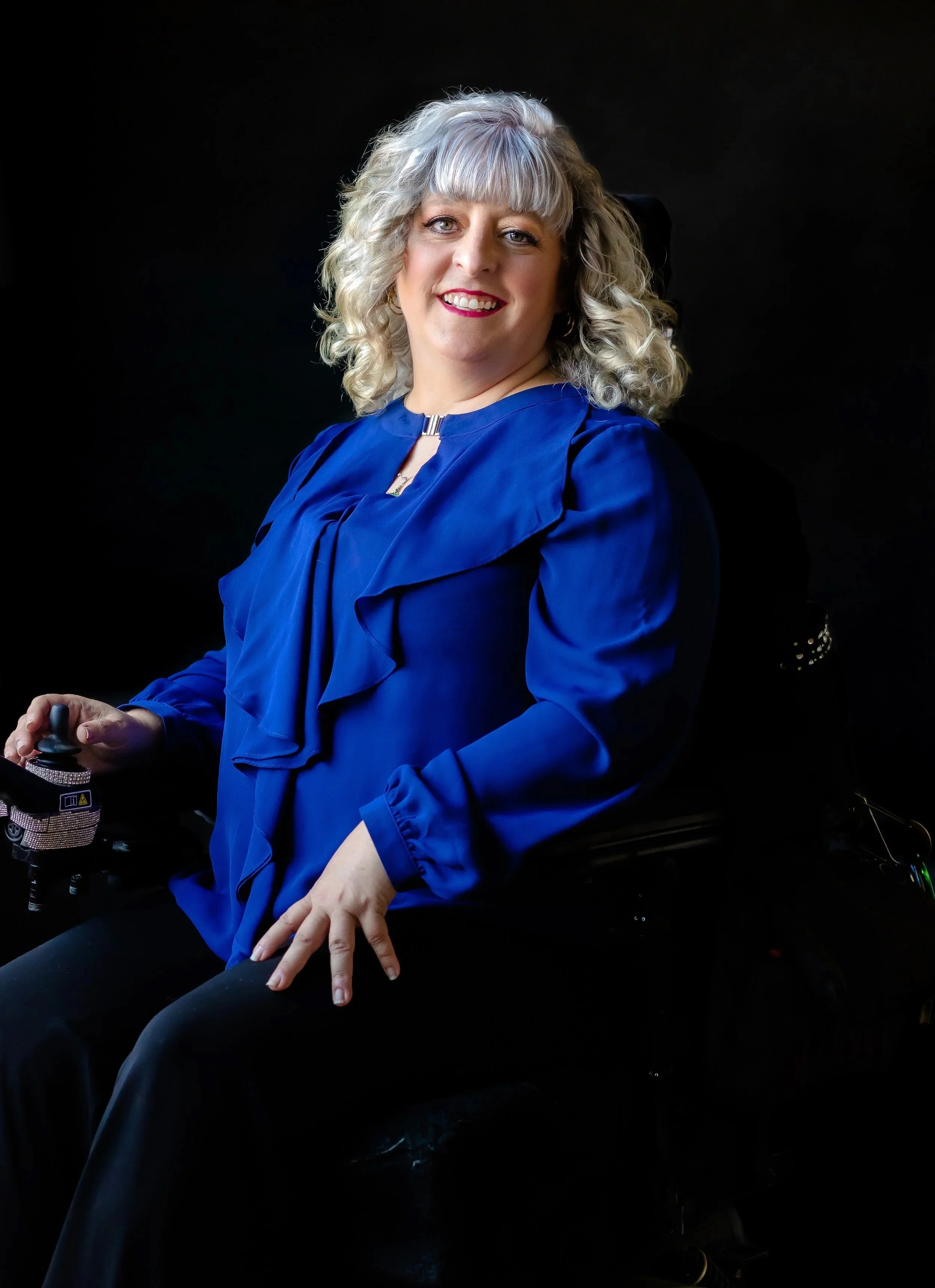 Woman with curly blonde hair wearing a blue blouse, sitting in a wheelchair, smiling at the camera.