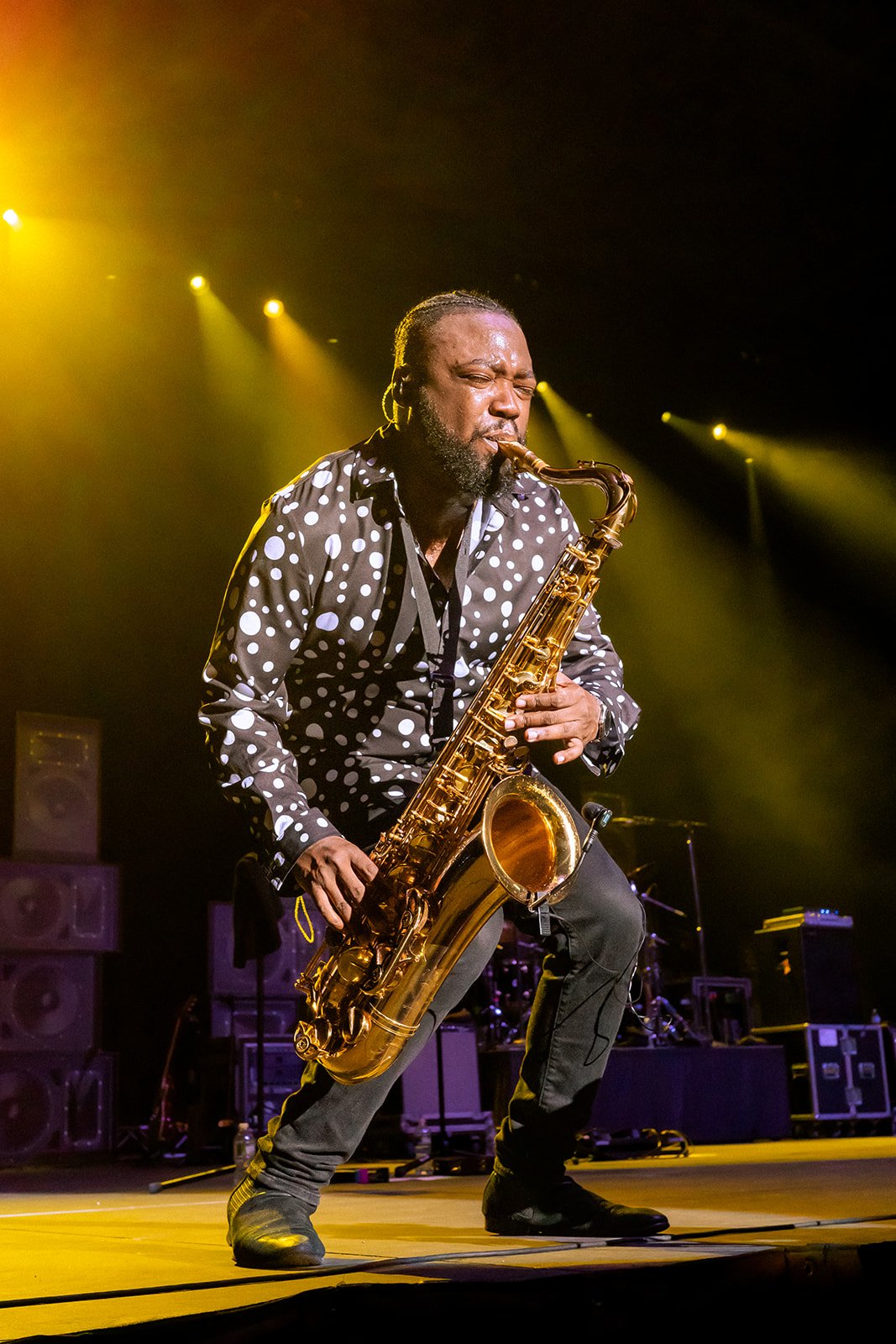 A man playing a saxophone on stage, wearing a black and white polka dot shirt and black pants amidst yellow stage lights.