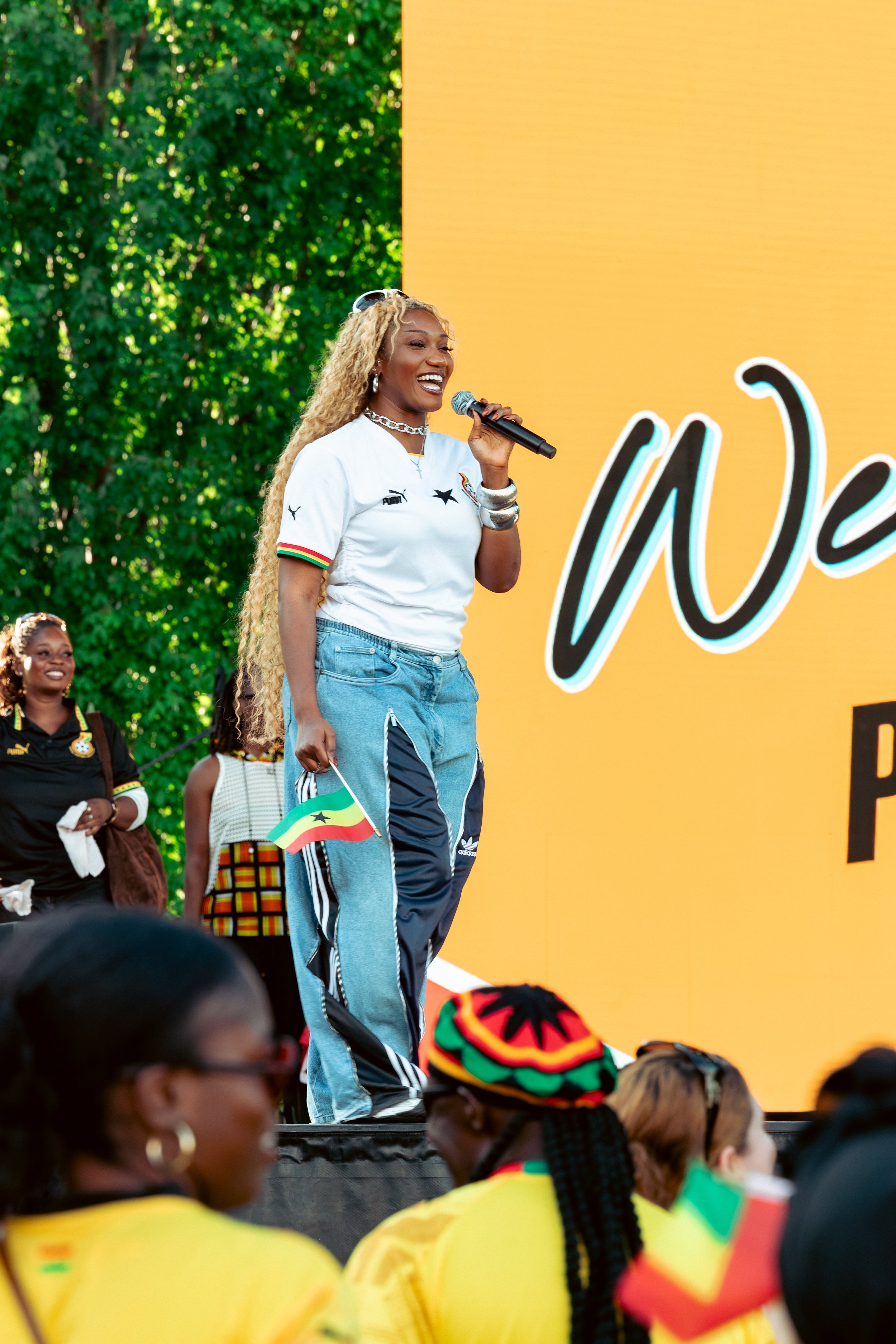 A woman with long curly blond hair, wearing a white sports shirt, blue track pants, and jewelry, is holding a microphone and speaking at an outdoor event. She is surrounded by a diverse group of people, some wearing colorful clothing and headwear, wi