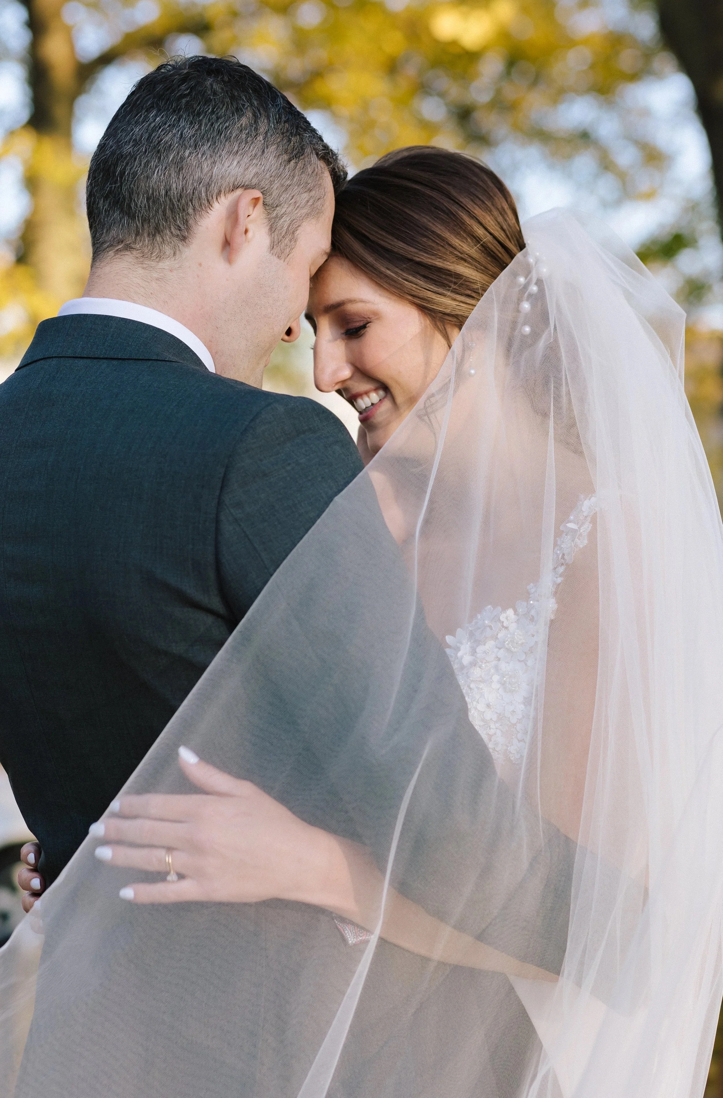 A bride and groom embrace outdoors, with their foreheads touching, smiling and smiling as their wedding veil drapes over them, with trees and sunlight in the background.