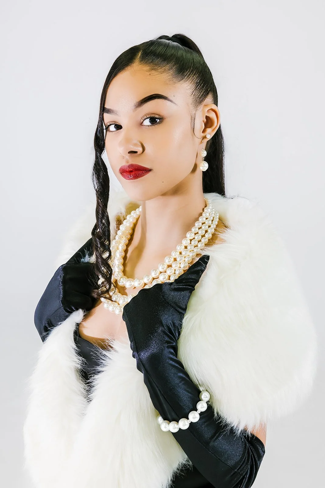 A woman wearing a white fur stole, black gloves with pearl embellishments, and pearl jewelry, posed against a plain white background.