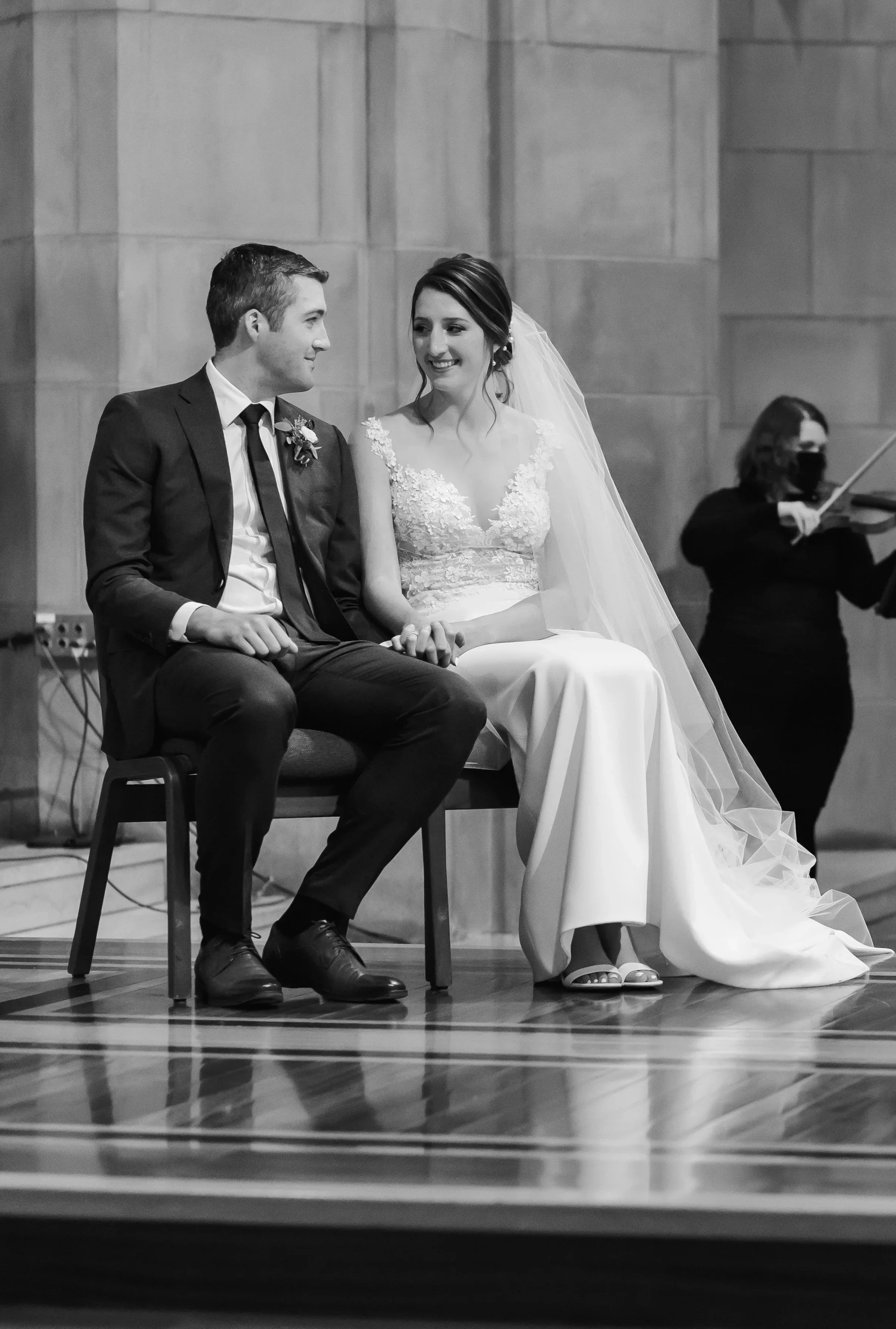 A black and white photo of a bride and groom sitting on a chair during their wedding ceremony. The bride is smiling and wearing a lace wedding gown with a veil, while the groom is dressed in a dark suit and tie. An orchestra musician, wearing a mask,