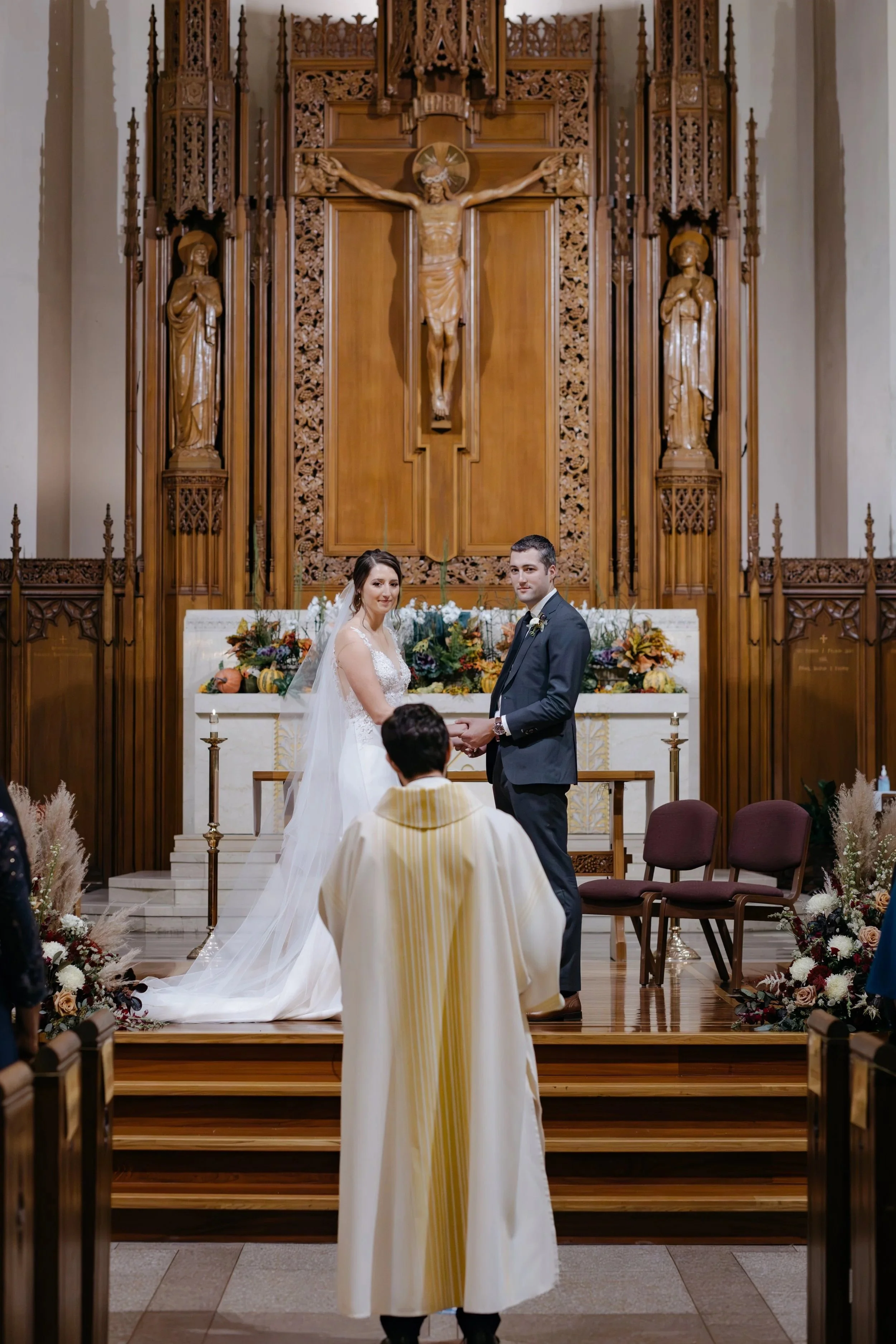A bride and groom holding hands during a wedding ceremony at the altar in a church. The altar is decorated with flowers and a large crucifix hanging on the wooden wall behind them. A priest in white robes is conducting the ceremony, and there are emp