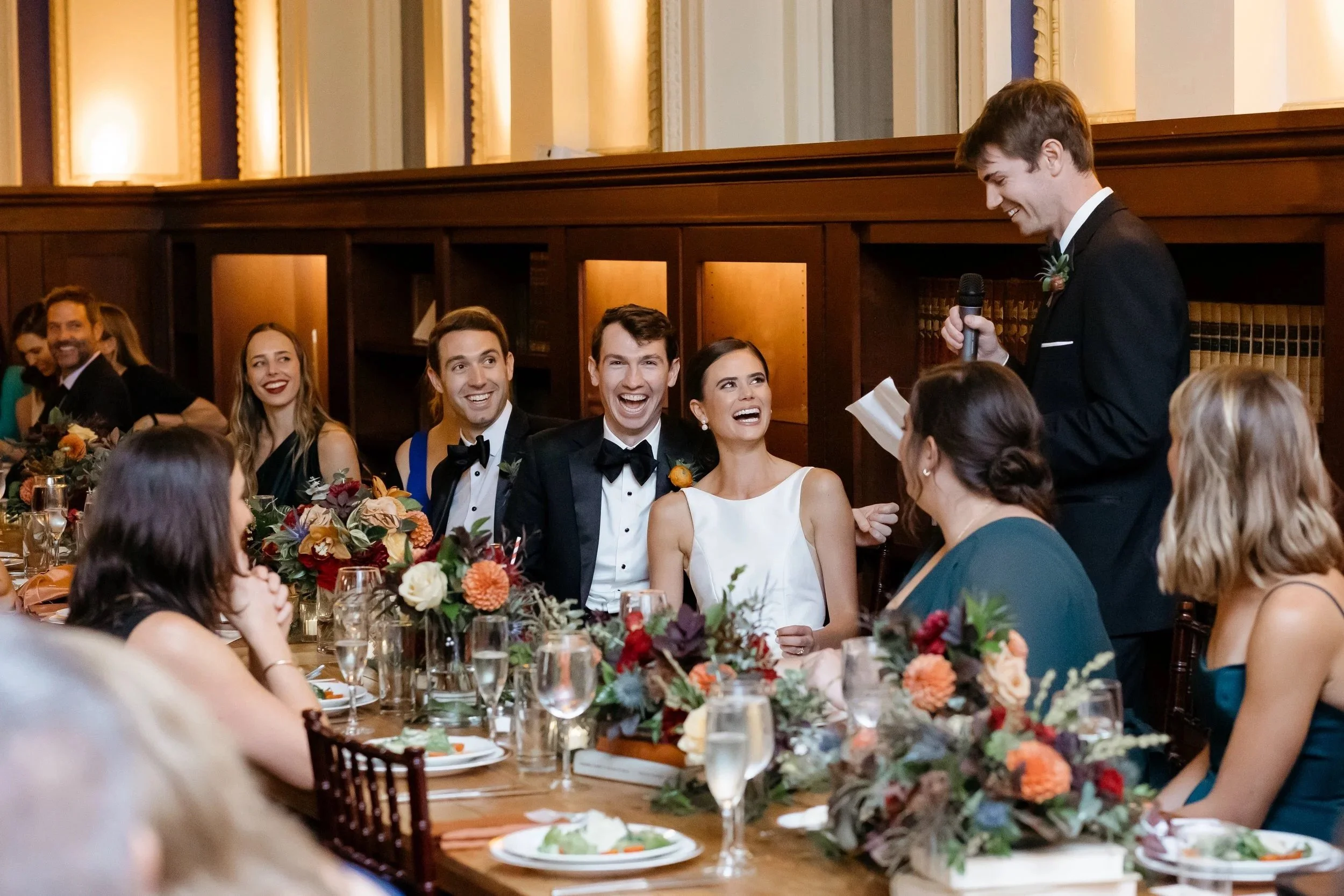 A wedding reception scene with the bride, groom, and guests listening to a speech. The groom and guests are smiling and laughing, with a man in the background holding a microphone and paper.