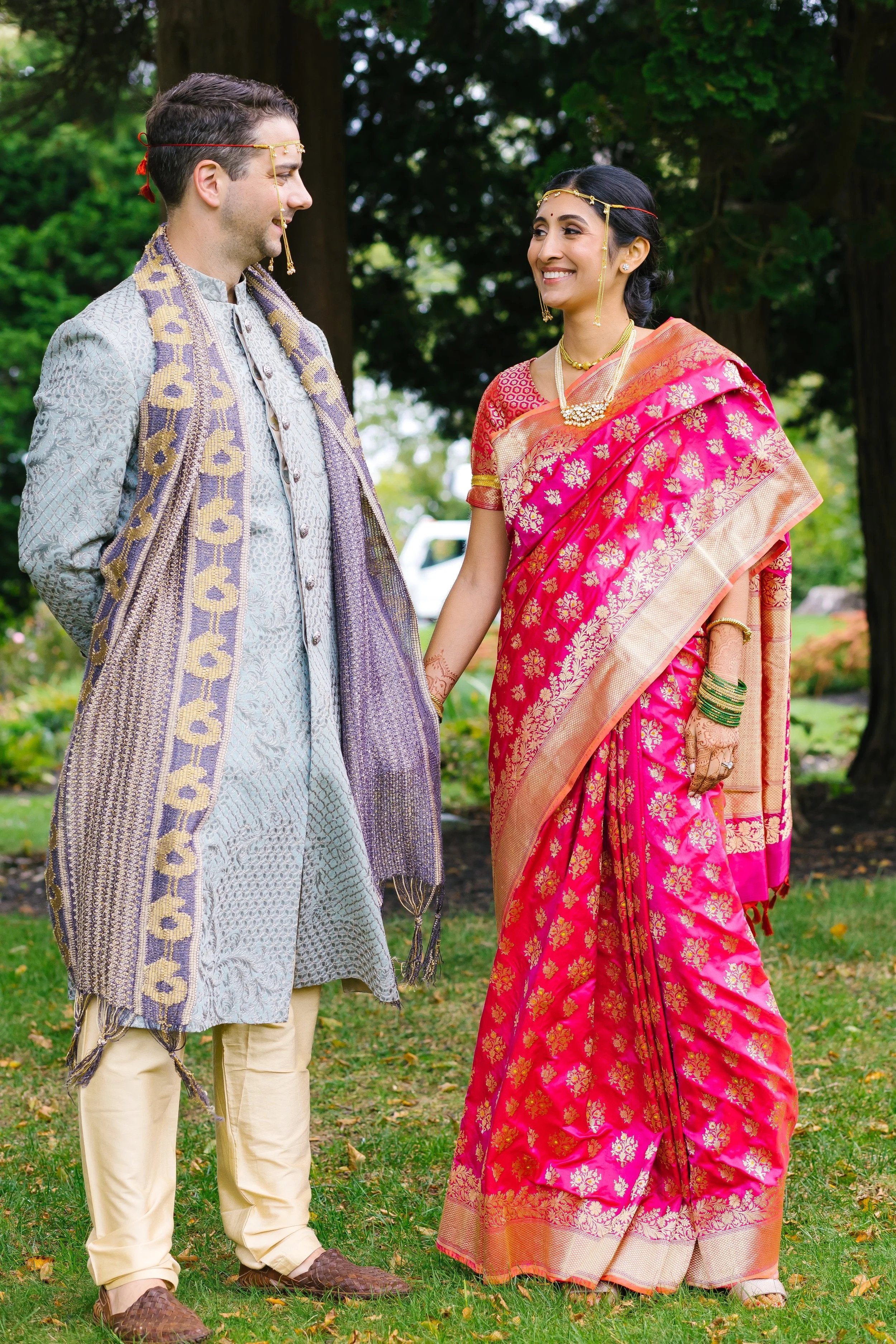 A couple in traditional Indian wedding attire holding hands outdoors, smiling at each other.