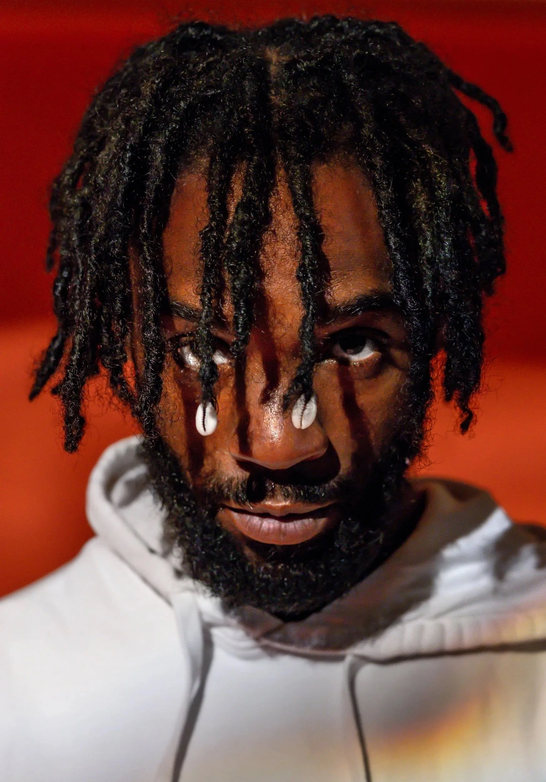 Close-up shot of a man with black dreadlocks, wearing a white hoodie, looking directly at the camera against a red background.