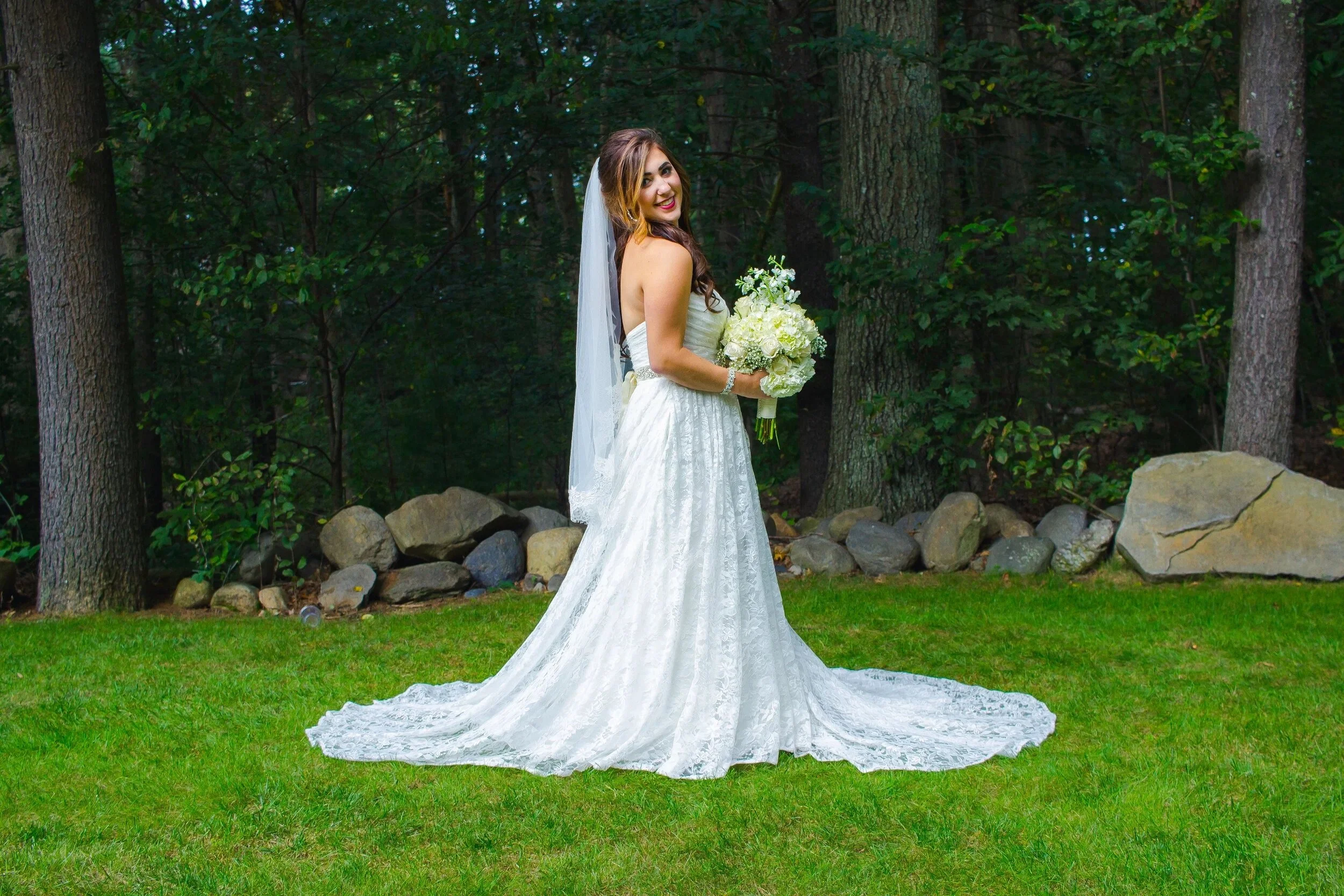 Bride in a strapless white wedding dress with a long train, holding a bouquet of white flowers, standing on a grassy area in a wooded outdoor setting.