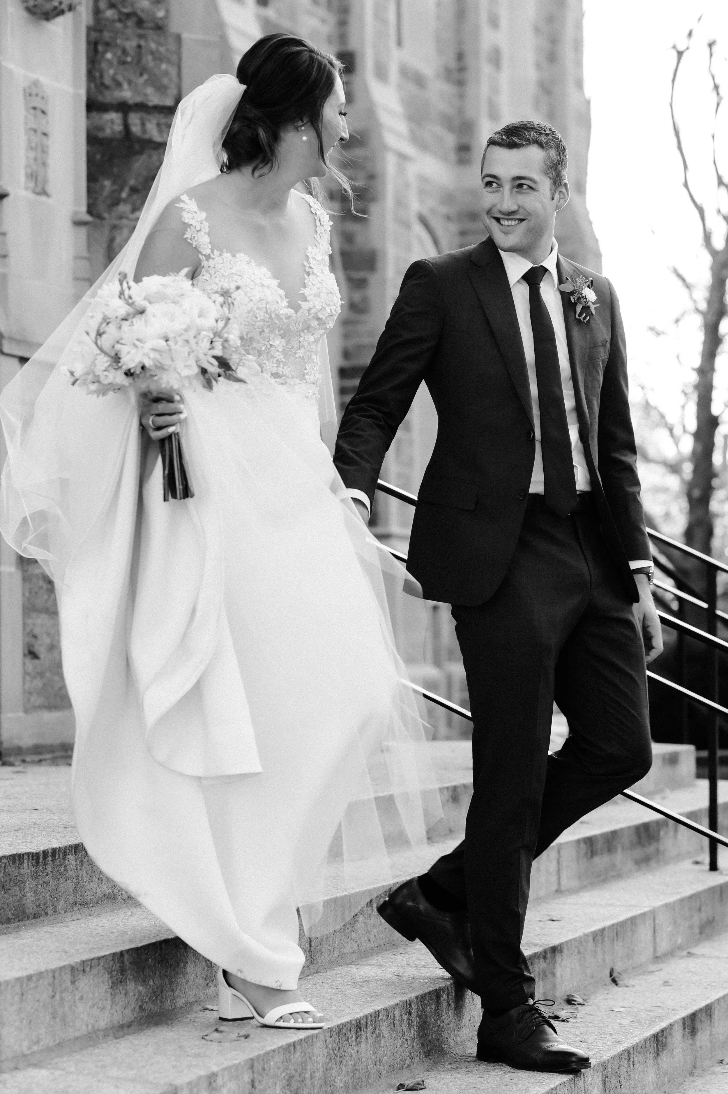 Black and white photo of a bride in a wedding gown and veil holding a bouquet, and a groom in a dark suit and tie holding her hand as they walk down stairs outside a church.