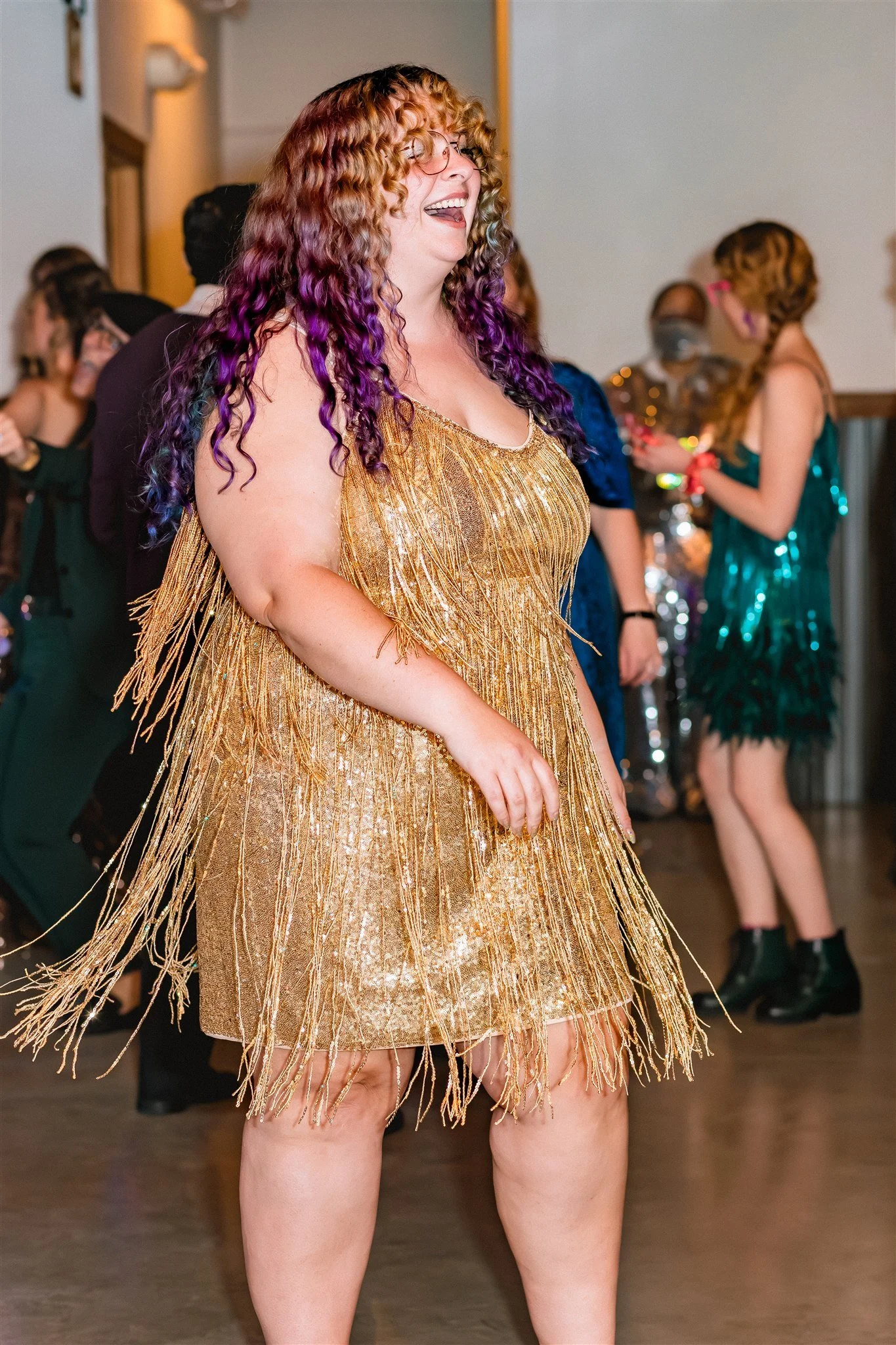 A woman with curly hair dyed purple and orange, laughing and dancing at a party, wearing a gold fringed dress.