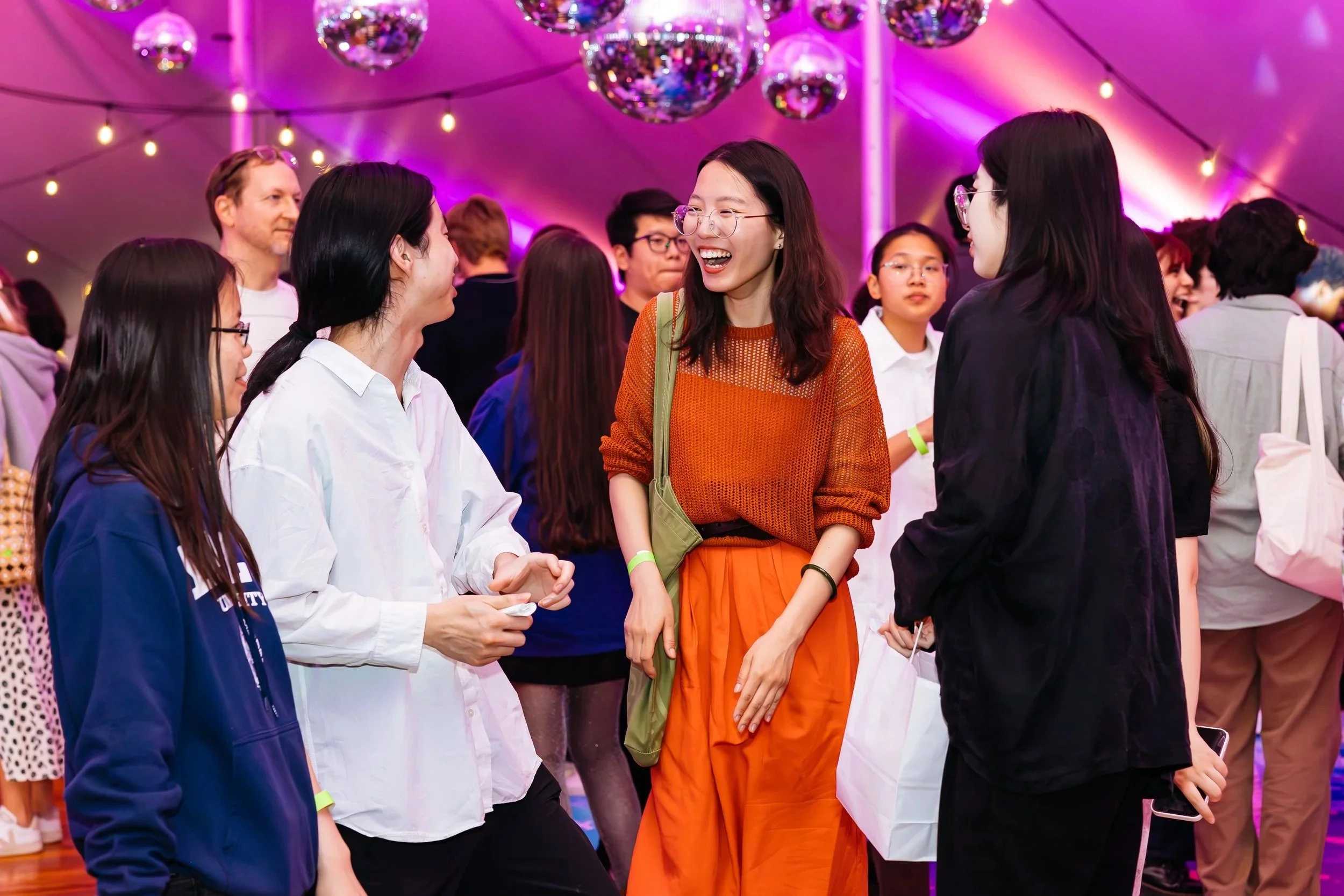 Group of diverse young adults at a lively party or social event, chatting and laughing under colorful lighting with disco balls overhead.