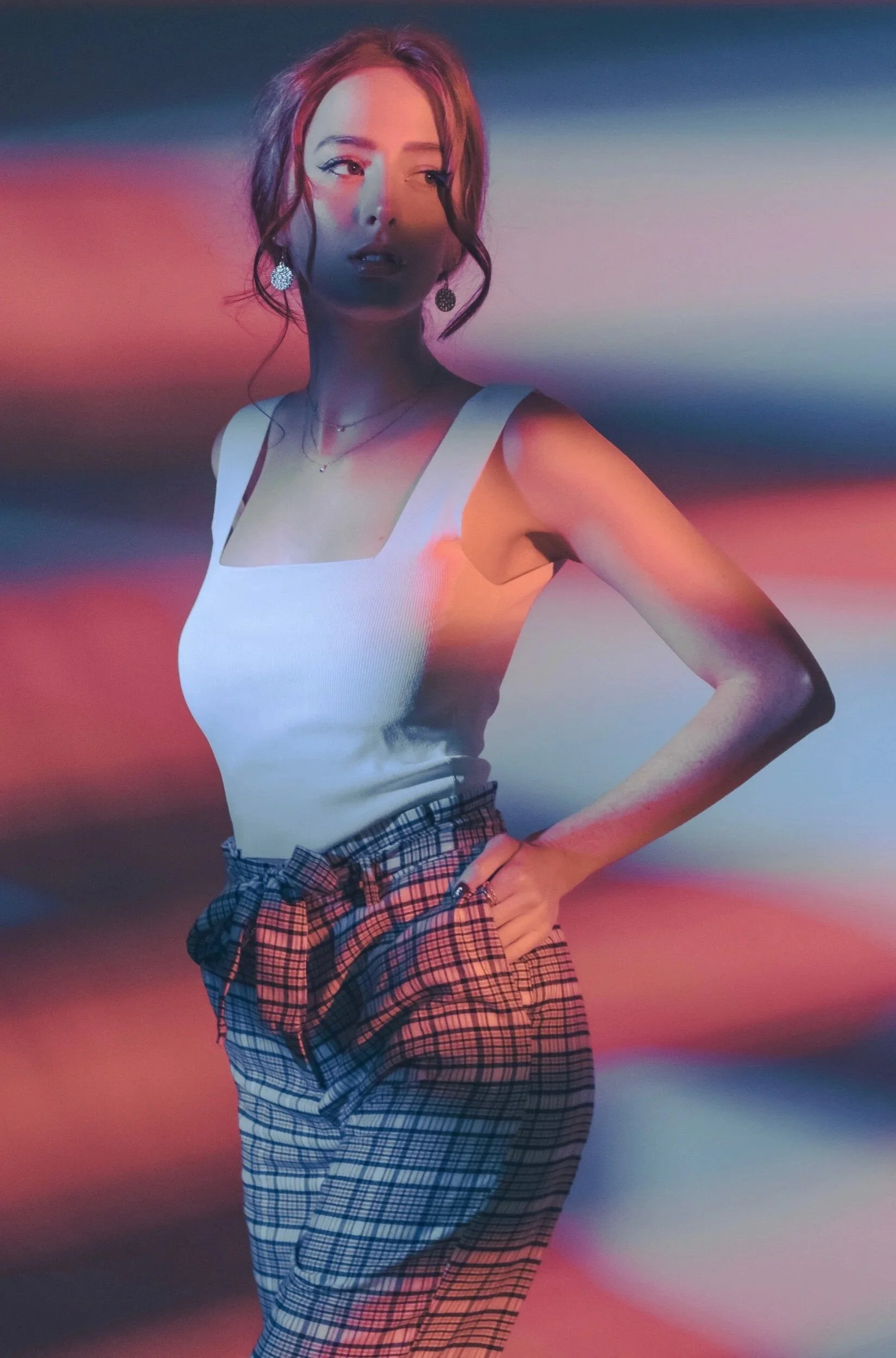 A young woman with brown hair styled in loose waves, wearing a white tank top with thick straps, plaid high-waisted pants, large earrings, and a necklace, standing with one hand on her hip against an abstract background with streaks of red, blue, and