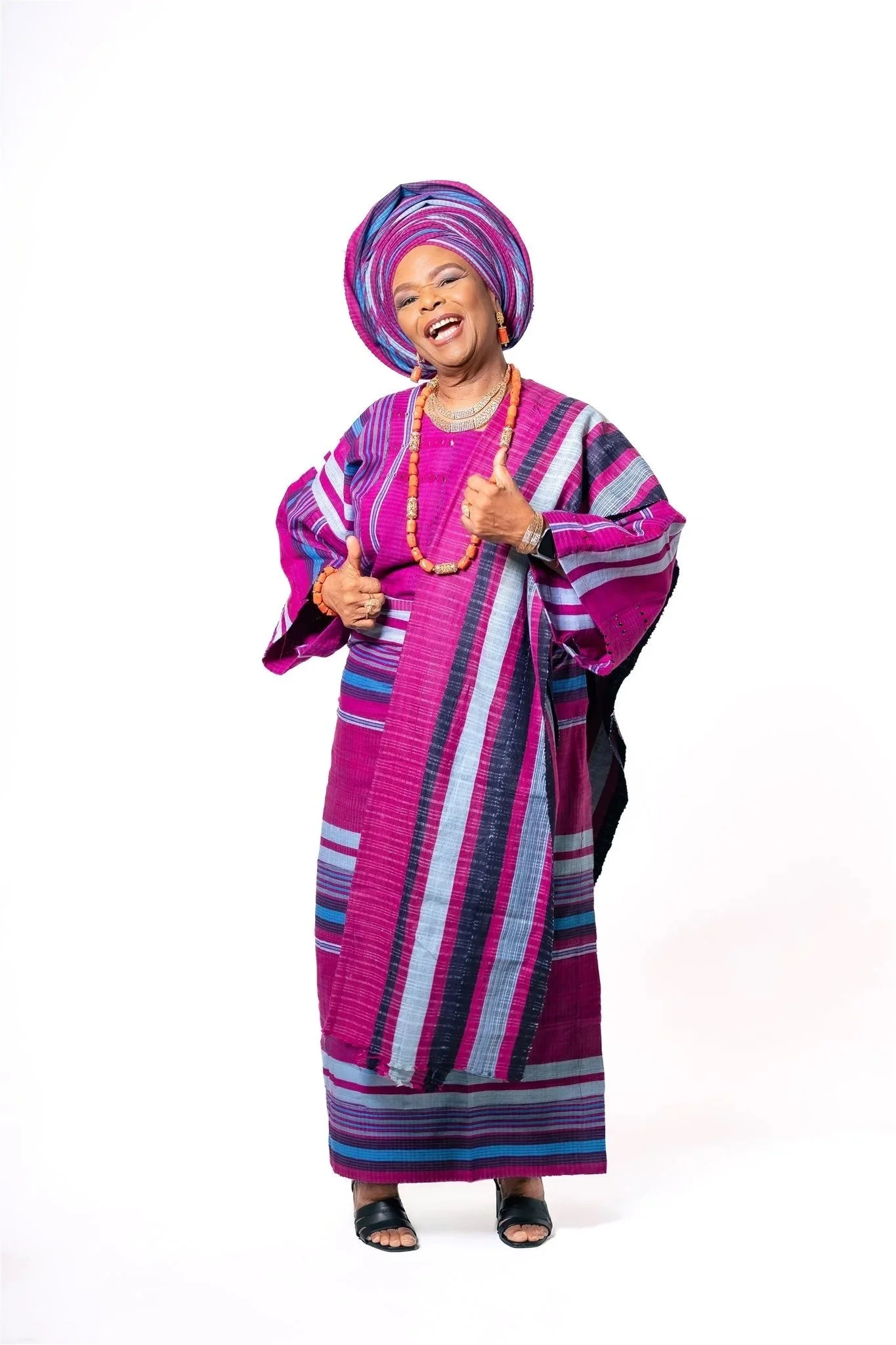 A smiling woman dressed in vibrant traditional African attire with a matching headwrap, necklace, and earrings, standing against a plain white background.