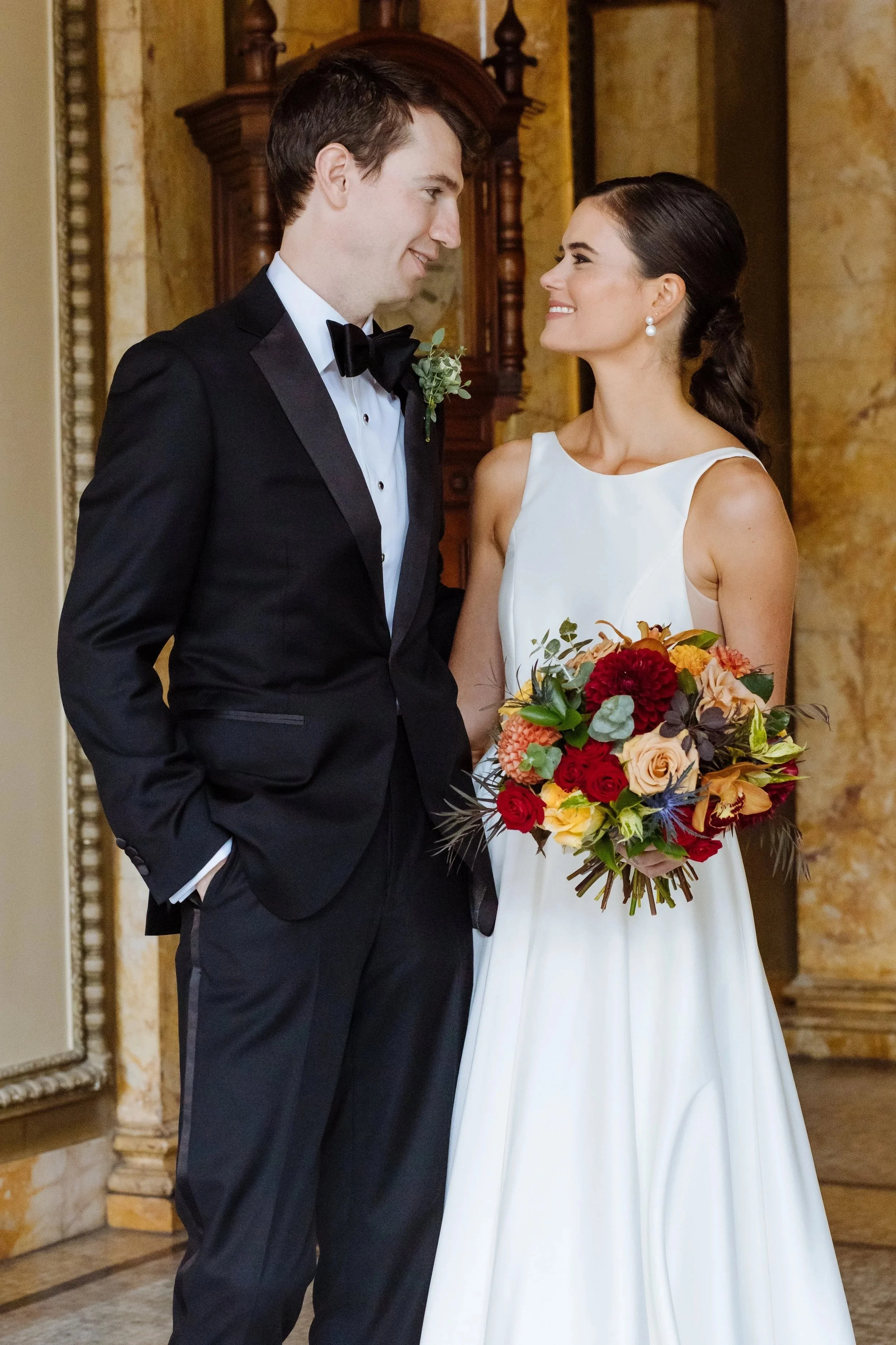 A bride and groom sharing a moment during their wedding, with the bride holding a colorful bouquet and smiling at the groom, dressed in a black tuxedo, in an elegant indoor setting.
