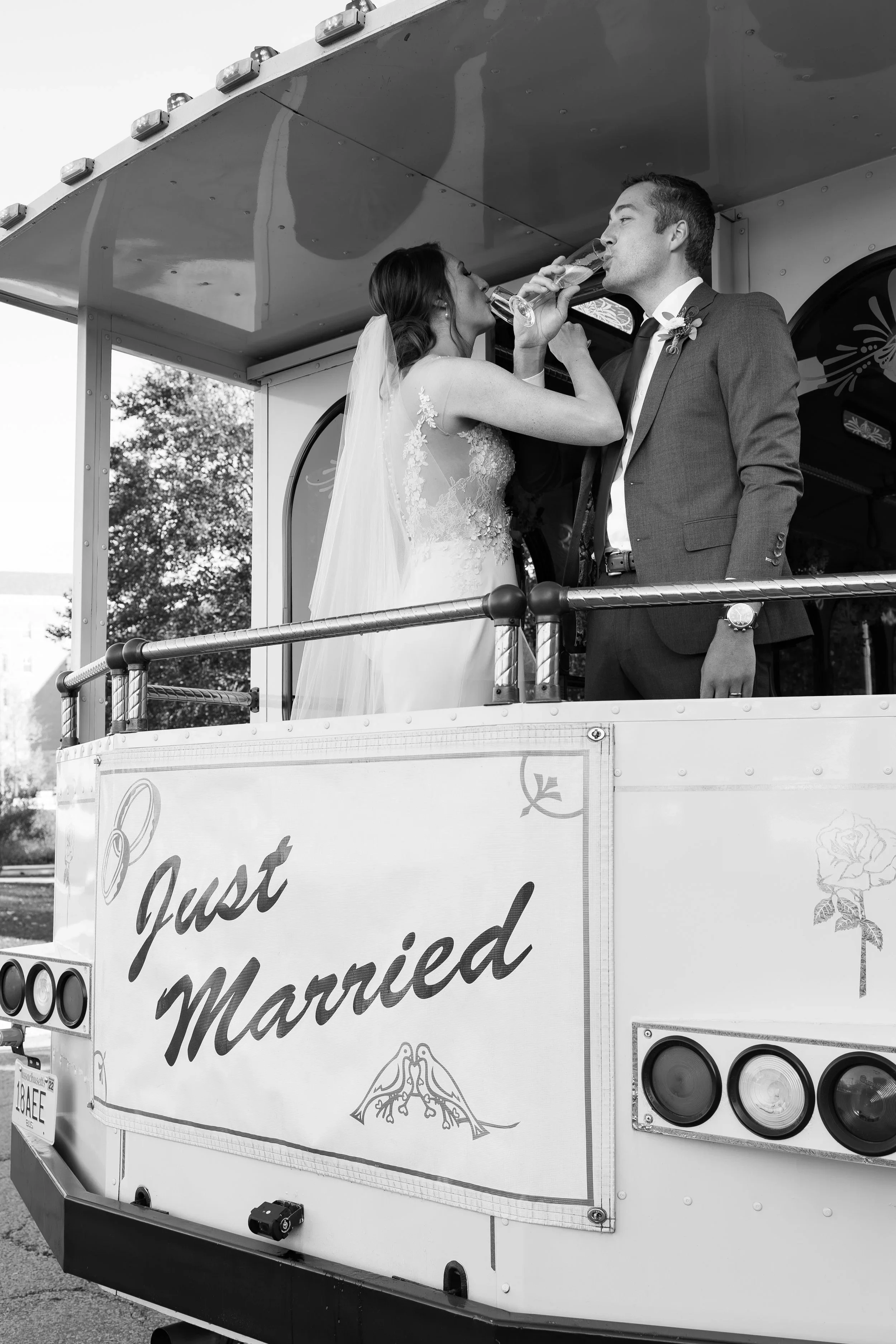 Black and white photo of a newlywed couple drinking champagne on a decorated tram with a "Just Married" sign.