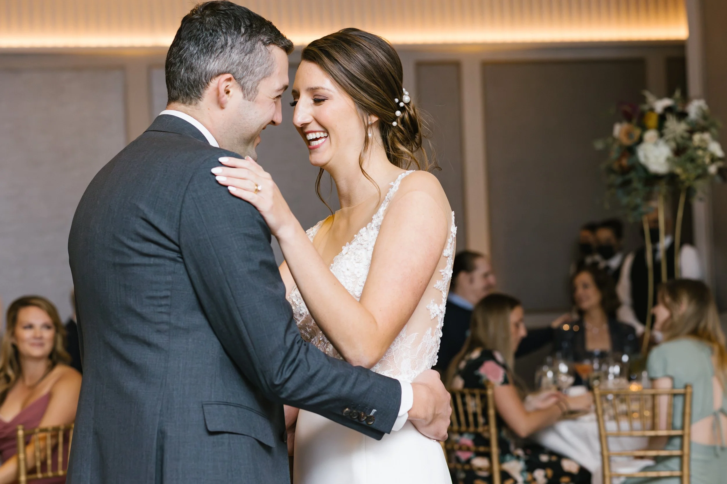 A bride and groom sharing a dance at their wedding reception, smiling and holding each other affectionately, with guests seated at tables in the background.
