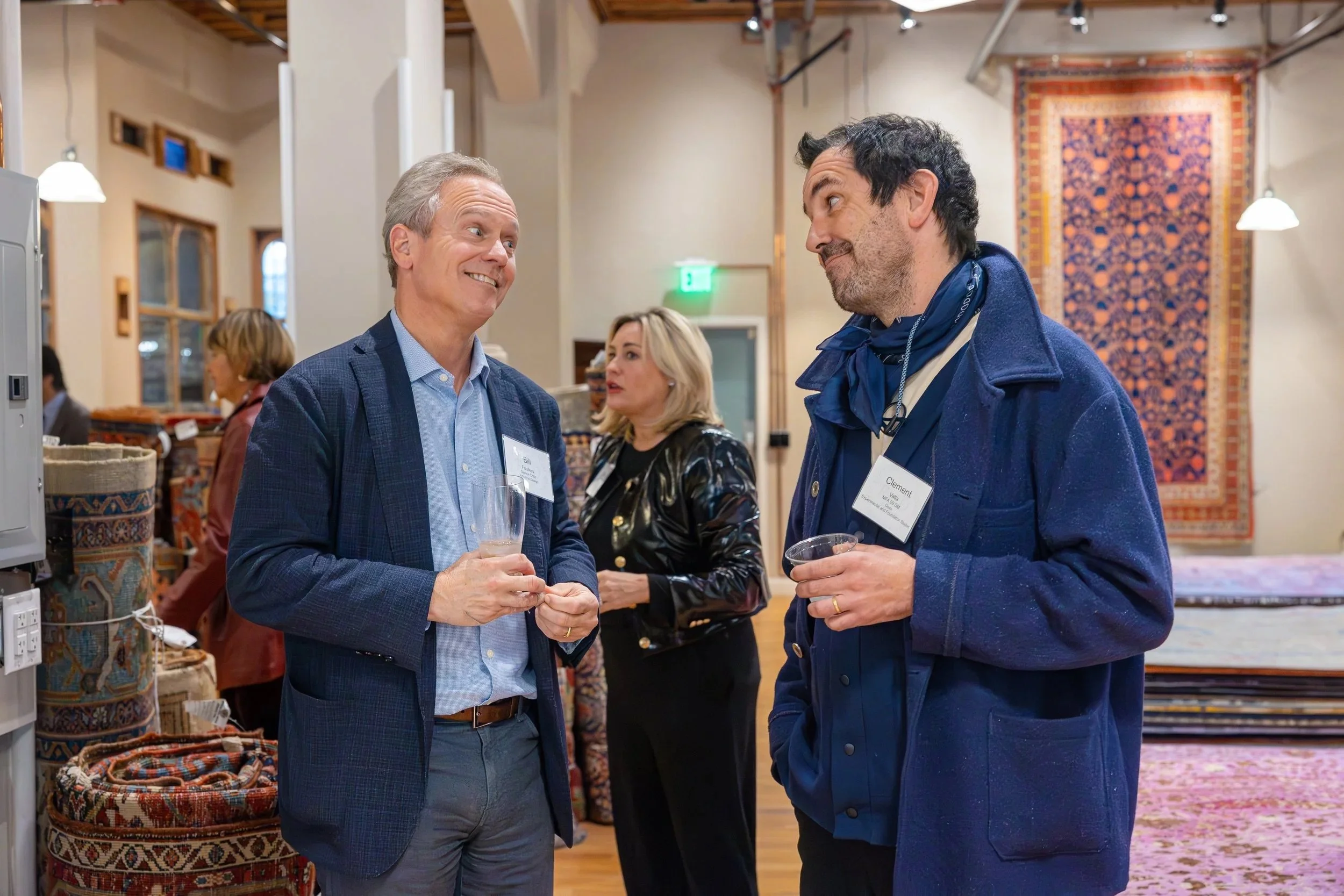 Two men engaged in conversation at a social gathering, holding drinks, in a room with stacked rugs and a decorative wall tapestry.