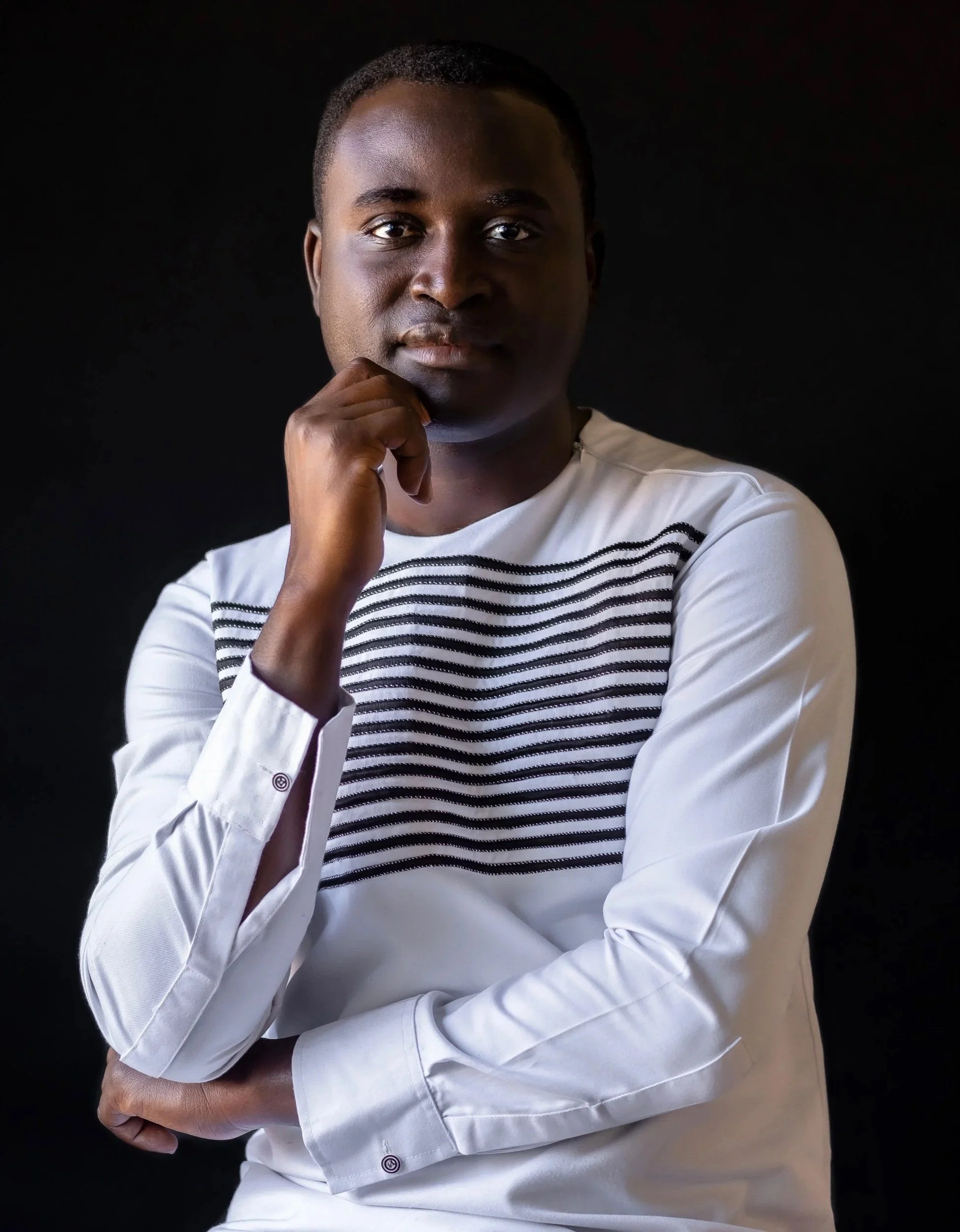 Portrait of a man wearing a white shirt with black stripes, standing against a black background, with his hand on his chin and looking at the camera.