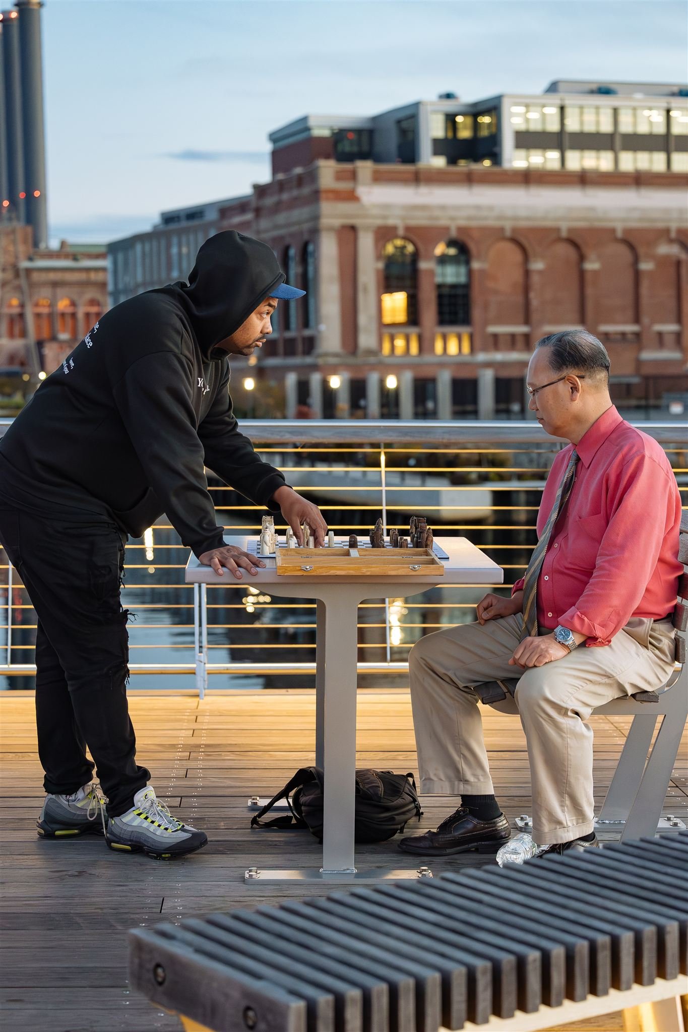 Two men playing chess outdoors at dusk, one standing and leaning over the chessboard, the other sitting on a bench.