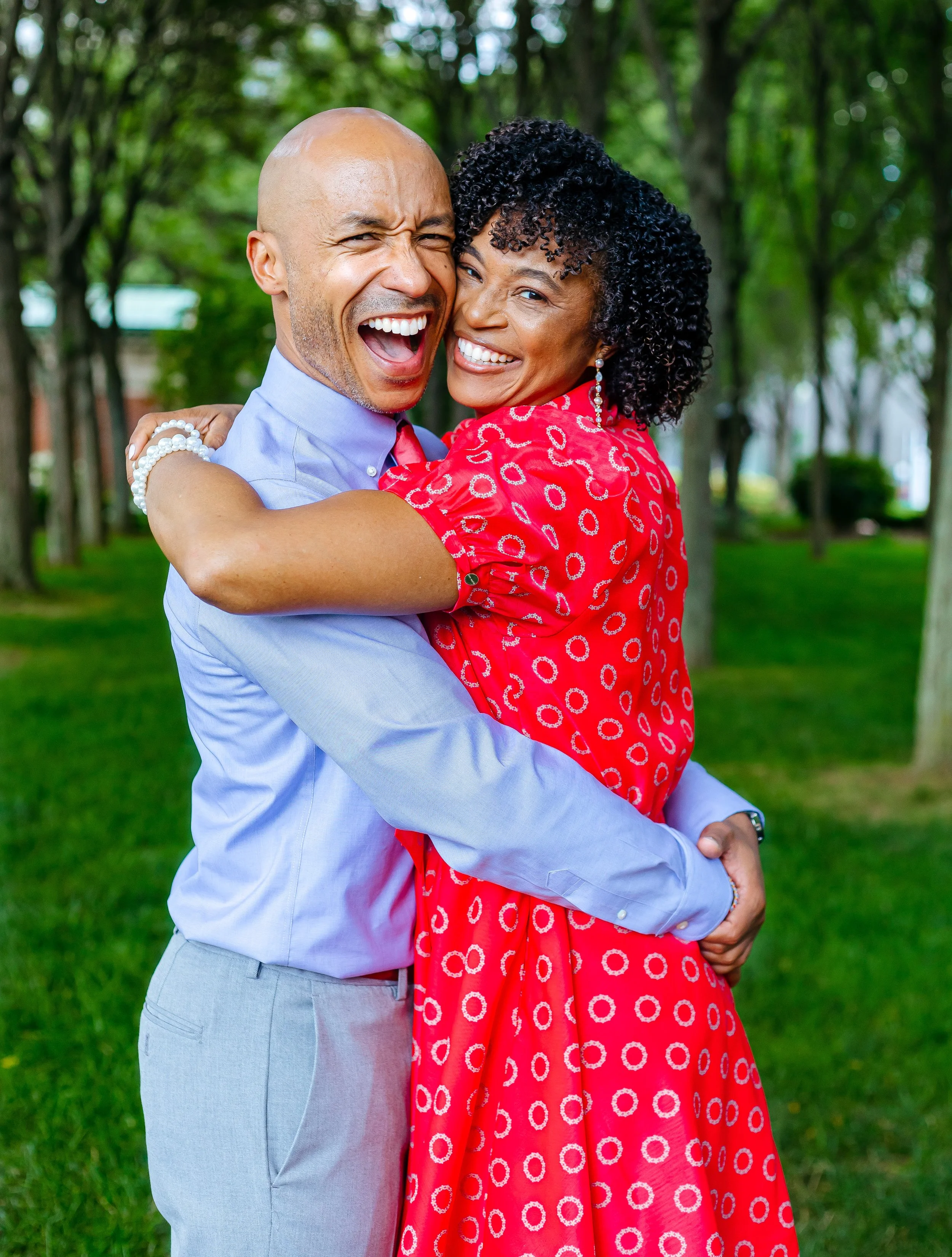 A happy couple hugging outdoors in a park with green trees and grass.