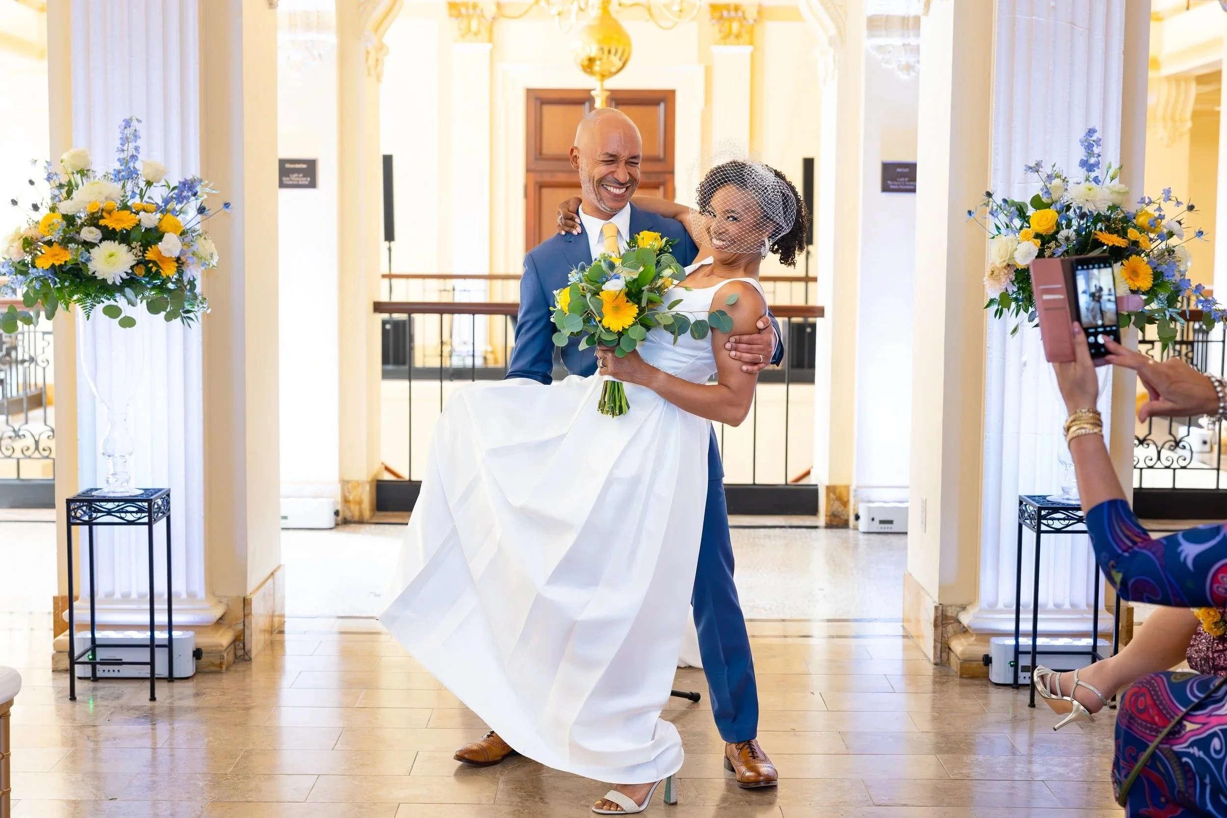 A happy bride and groom smiling, with the groom lifting the bride while she holds a bouquet of yellow and white flowers, at their wedding ceremony. A woman is taking a photo in the background.