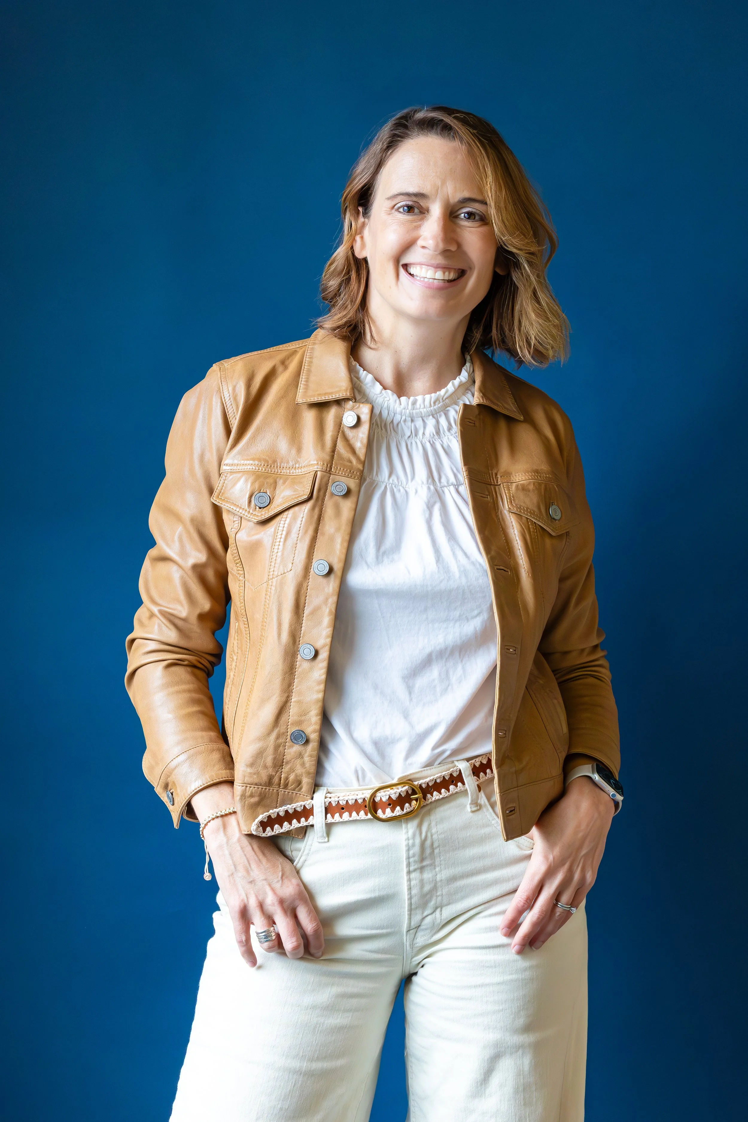 A woman with shoulder-length brown hair smiling, wearing a light brown leather jacket over a white ruffled blouse, cream-colored pants, and a patterned belt, posed against a blue background.