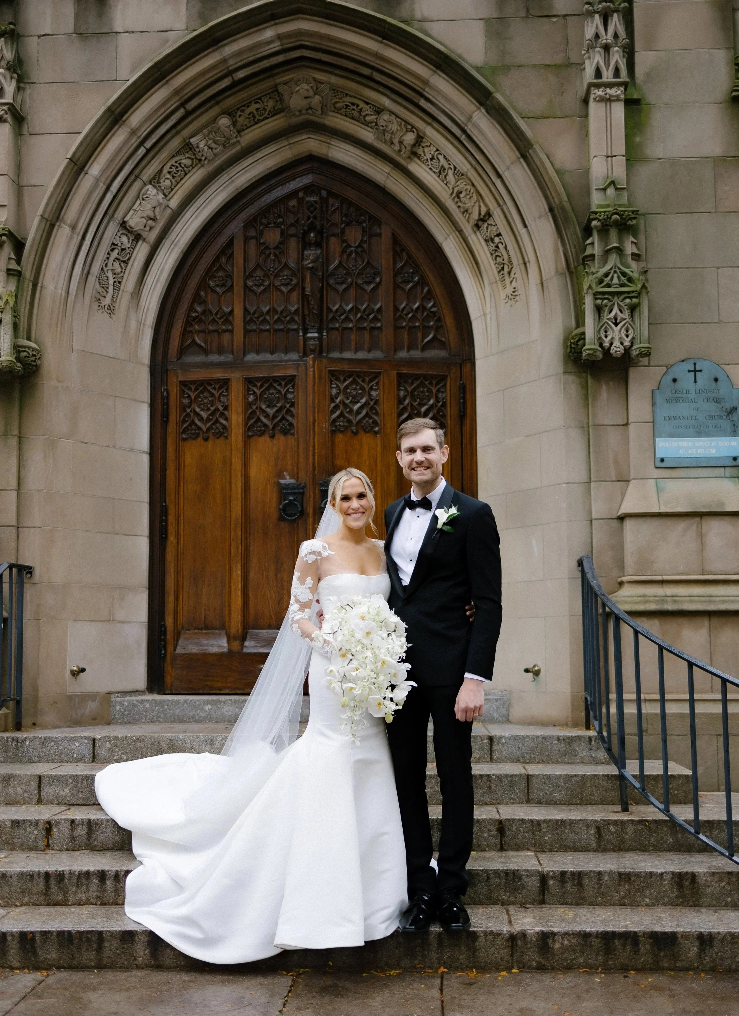 A bride and groom standing on church steps, smiling, with the bride holding a large white floral bouquet, outside a historic church with an ornate wooden door.