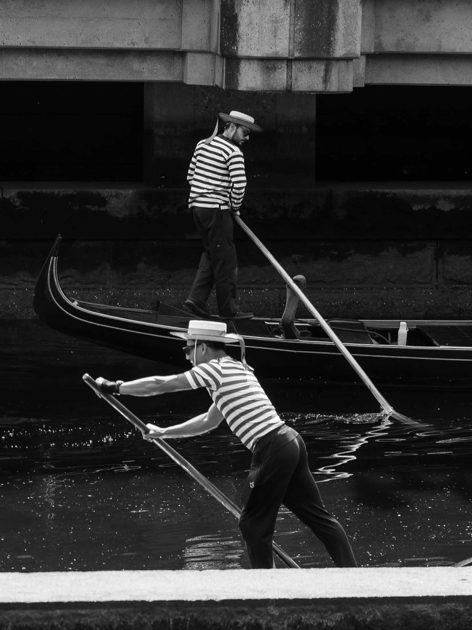 Two people wearing striped shirts and wide-brimmed hats, riding on a gondola and standing on a dock in a canal, with a bridge overhead.