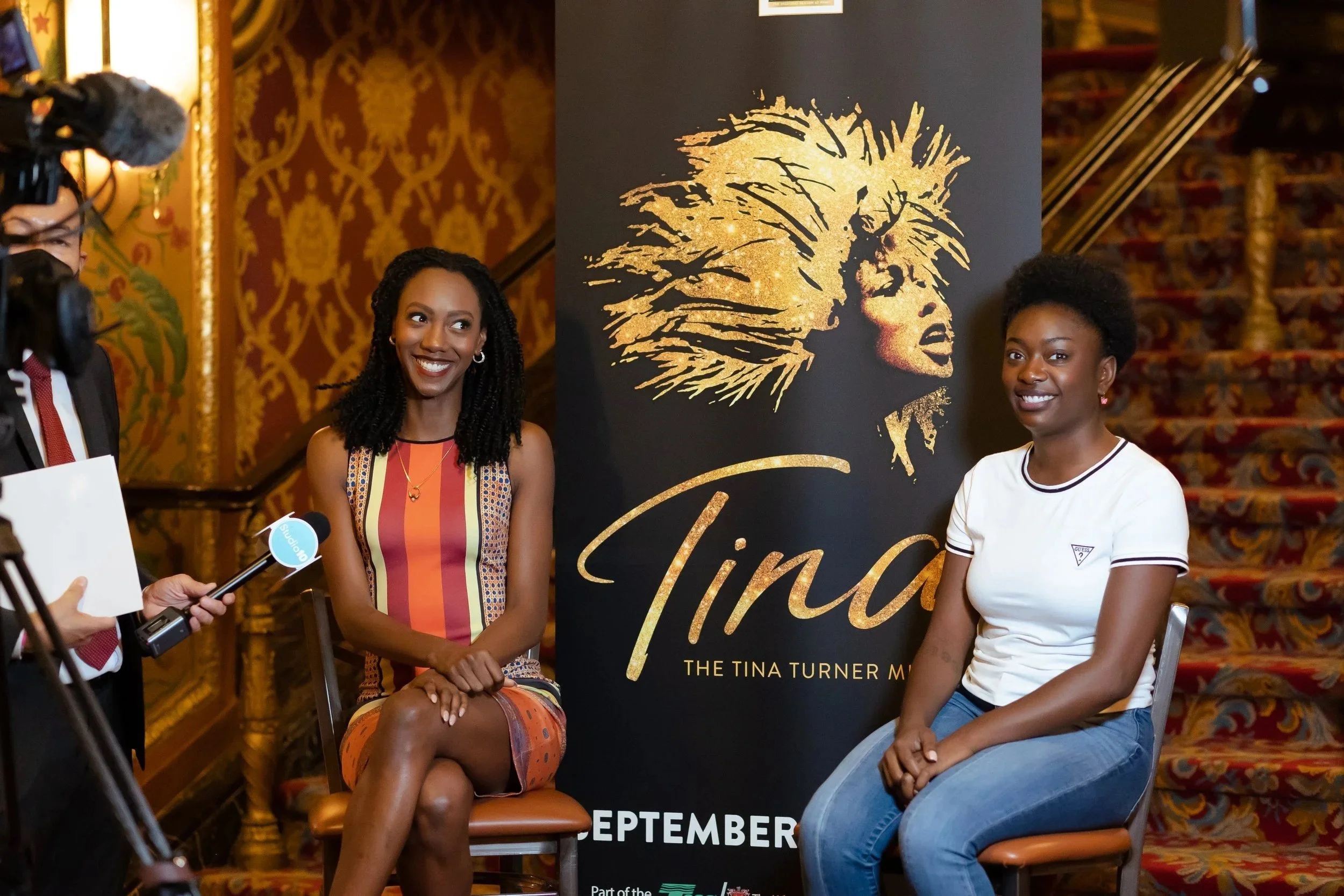 Two women being interviewed in front of a poster for 'Tina: The Tina Turner Musical'. They are sitting on chairs in a fancy, ornately decorated room with staircase in the background.