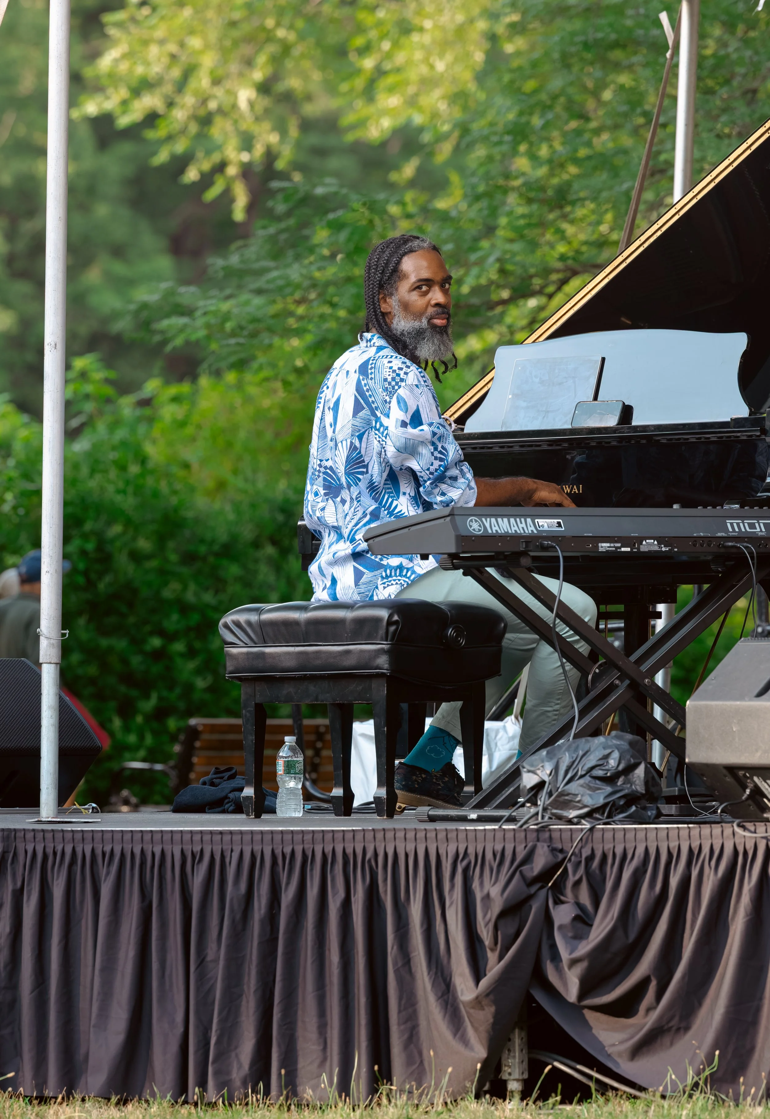 A man playing a grand piano and a keyboard on an outdoor stage surrounded by green trees.