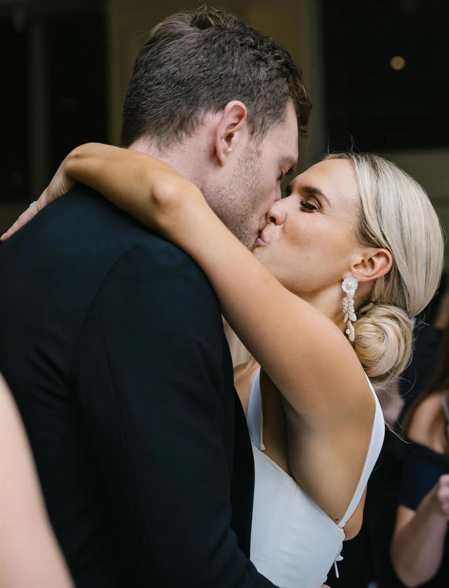 A couple is kissing closely, with the bride wrapping her arms around her husband's shoulders, both with eyes closed.