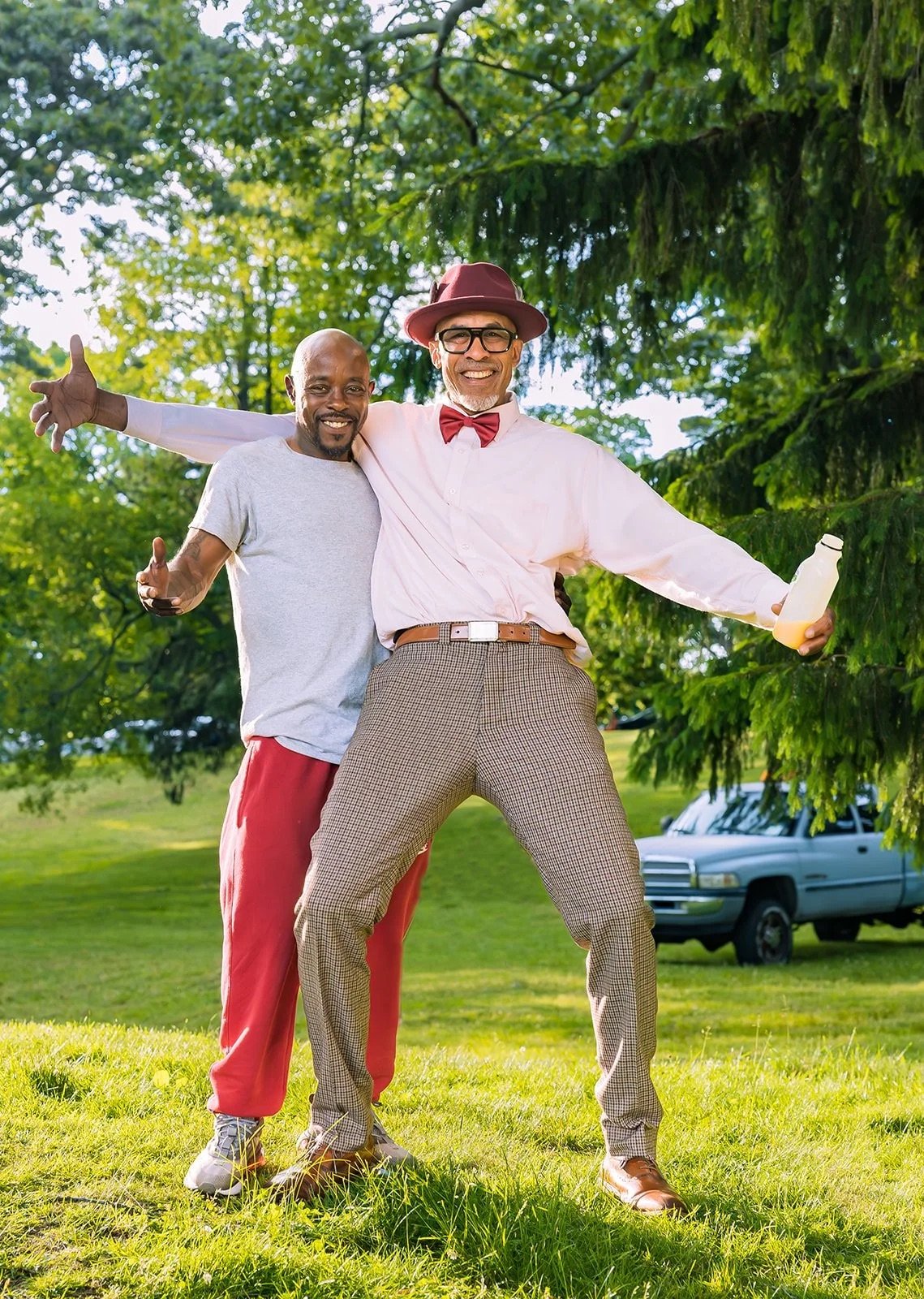Two men smiling and embracing outdoors on a sunny day, with trees and a parked car in the background. One man wears casual clothing, and the other is dressed in vintage style with a hat, glasses, bowtie, and checkered trousers.