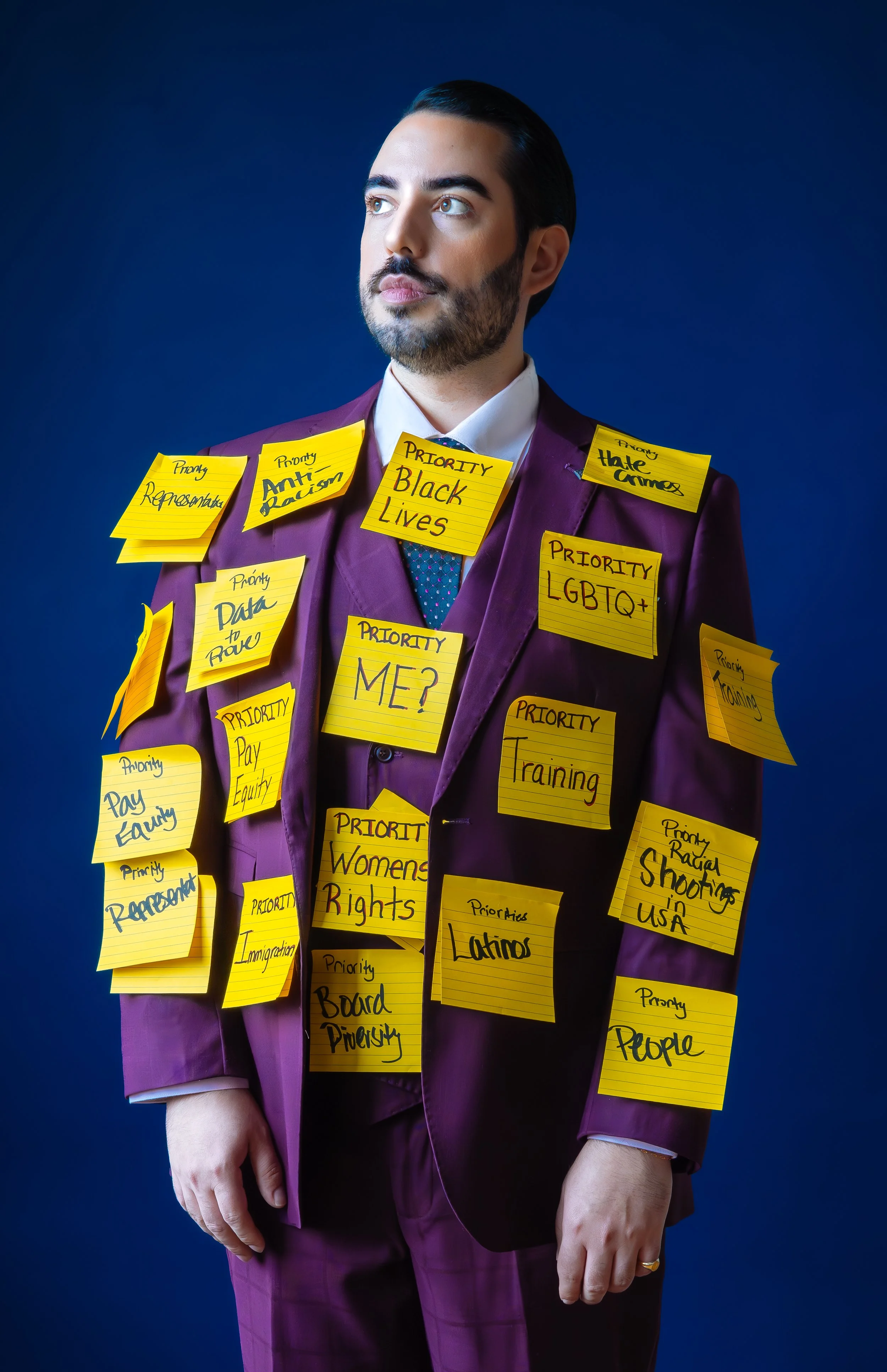 A man in a purple suit with dark hair and a beard, looking to the side, is covered with yellow sticky notes with various social justice and political messages written on them, such as "Black Lives," "Women Rights," and "LGBTQ+."