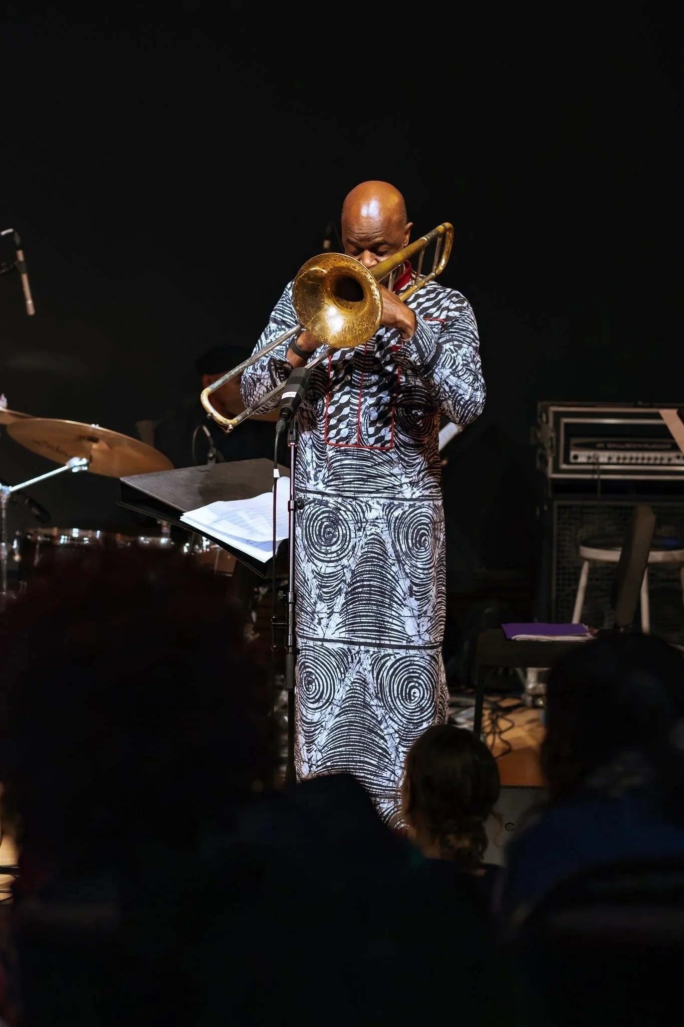 A man playing a trumpet on stage, wearing a patterned outfit, with music stands and musical equipment around him, and an audience watching.