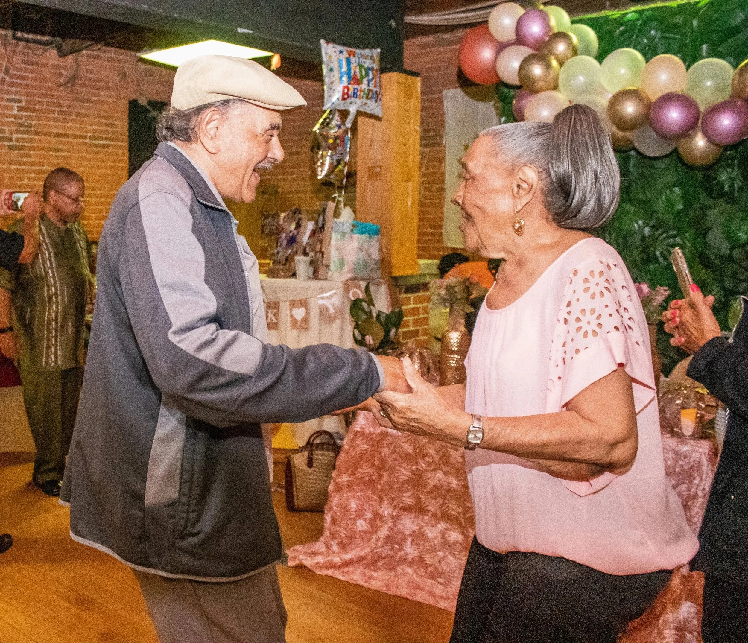 An elderly man and woman are smiling and holding hands while dancing at a birthday celebration with balloons and decorations.