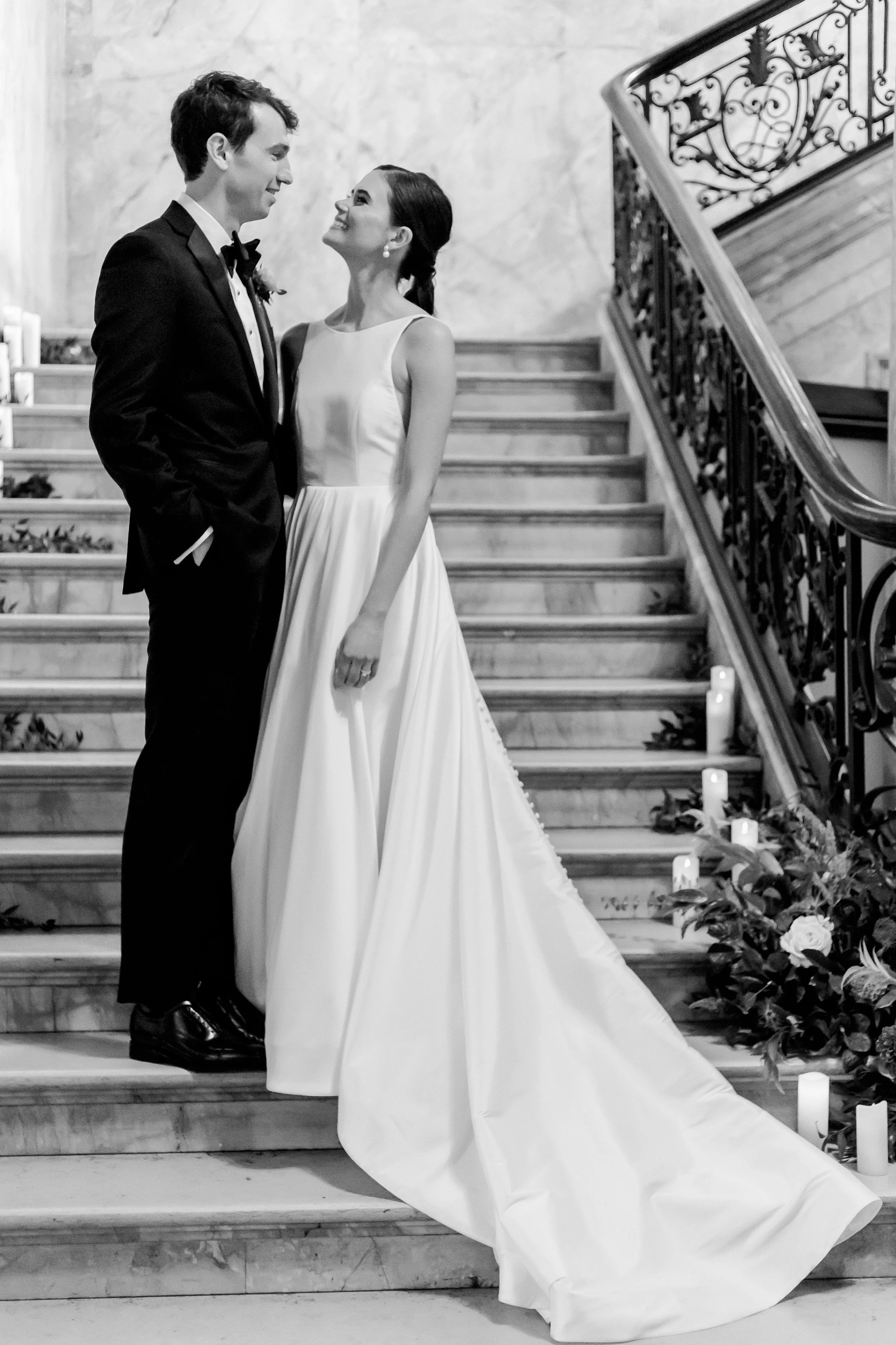 A bride and groom on a staircase, gazing at each other, in a black and white wedding photo. The bride wears a long, elegant white gown, and the groom a classic black tuxedo.