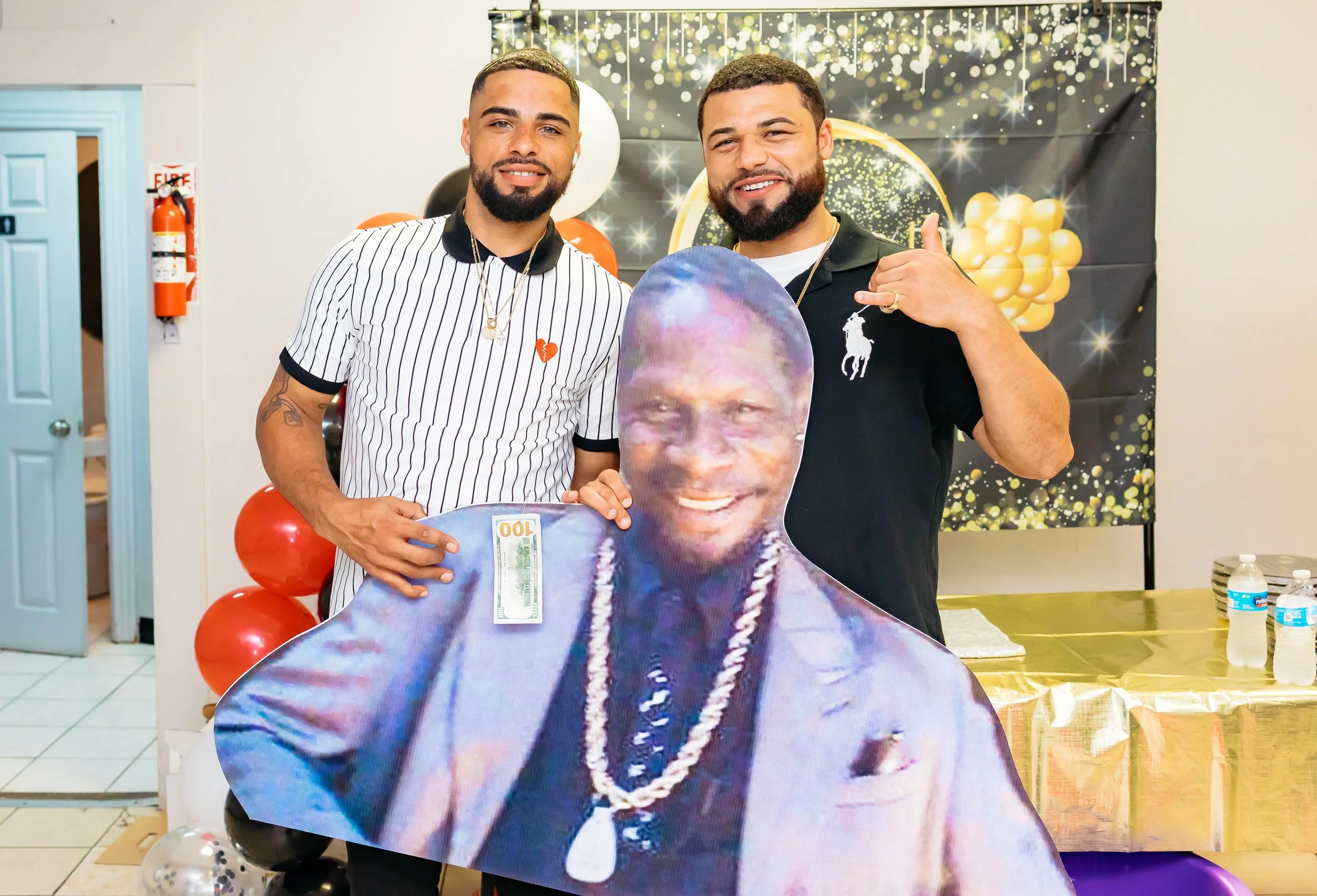 Two men celebrating at a party, one holding a large cutout of a man's face with a 100-dollar bill attached, gold and black themed decorations behind them, and balloons in the background.