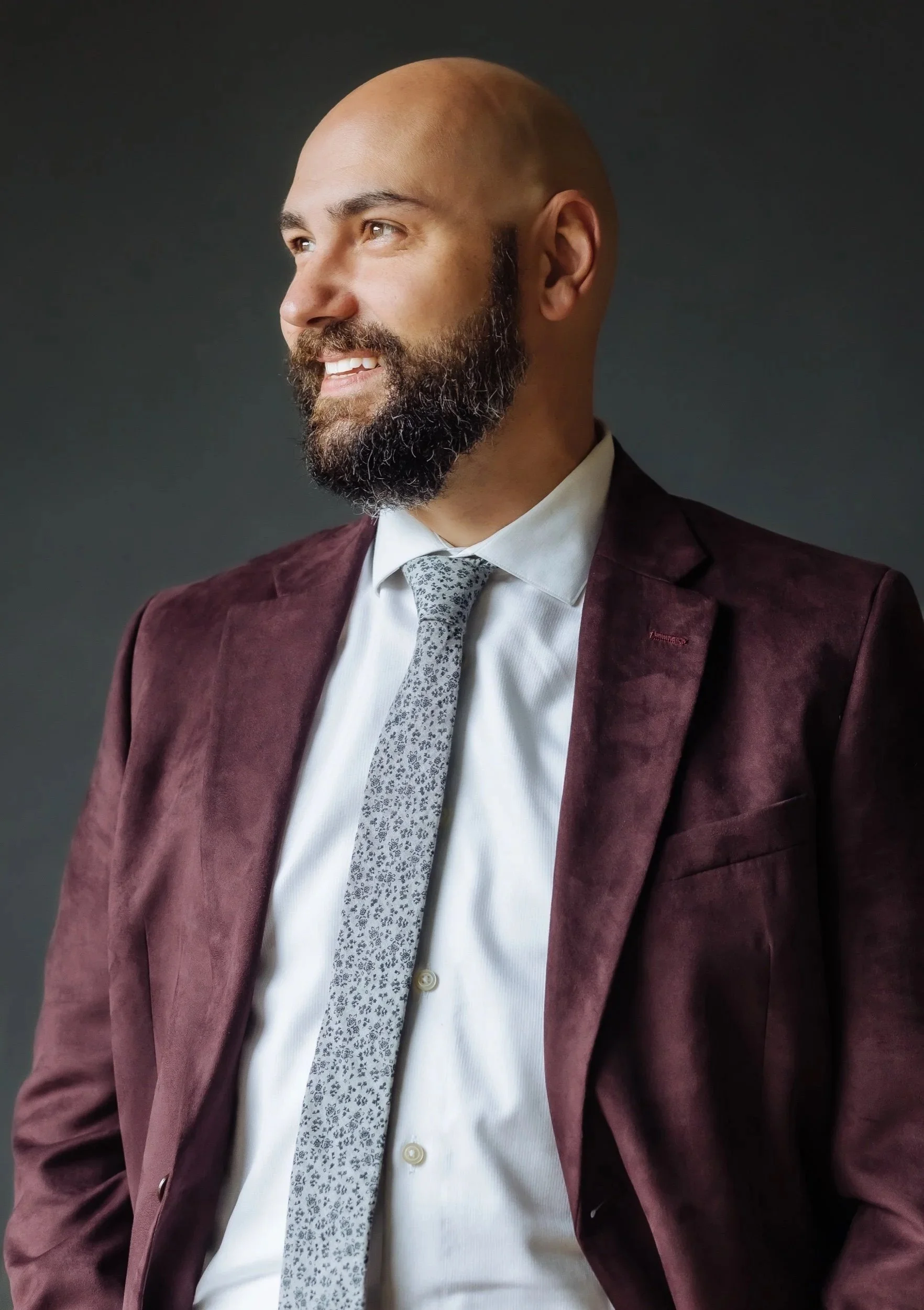 A man with a beard and bald head wearing a white shirt, gray patterned tie, and maroon blazer, smiling and looking to the side against a dark background.