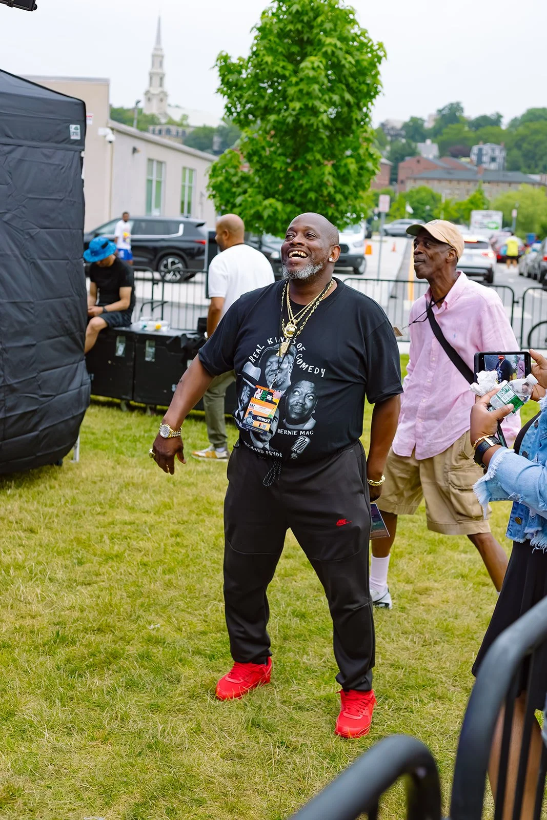 A man smiling and wearing a black t-shirt with pictures of Bernie Mac, gold chains, black pants, and red shoes at an outdoor event with other people in the background.