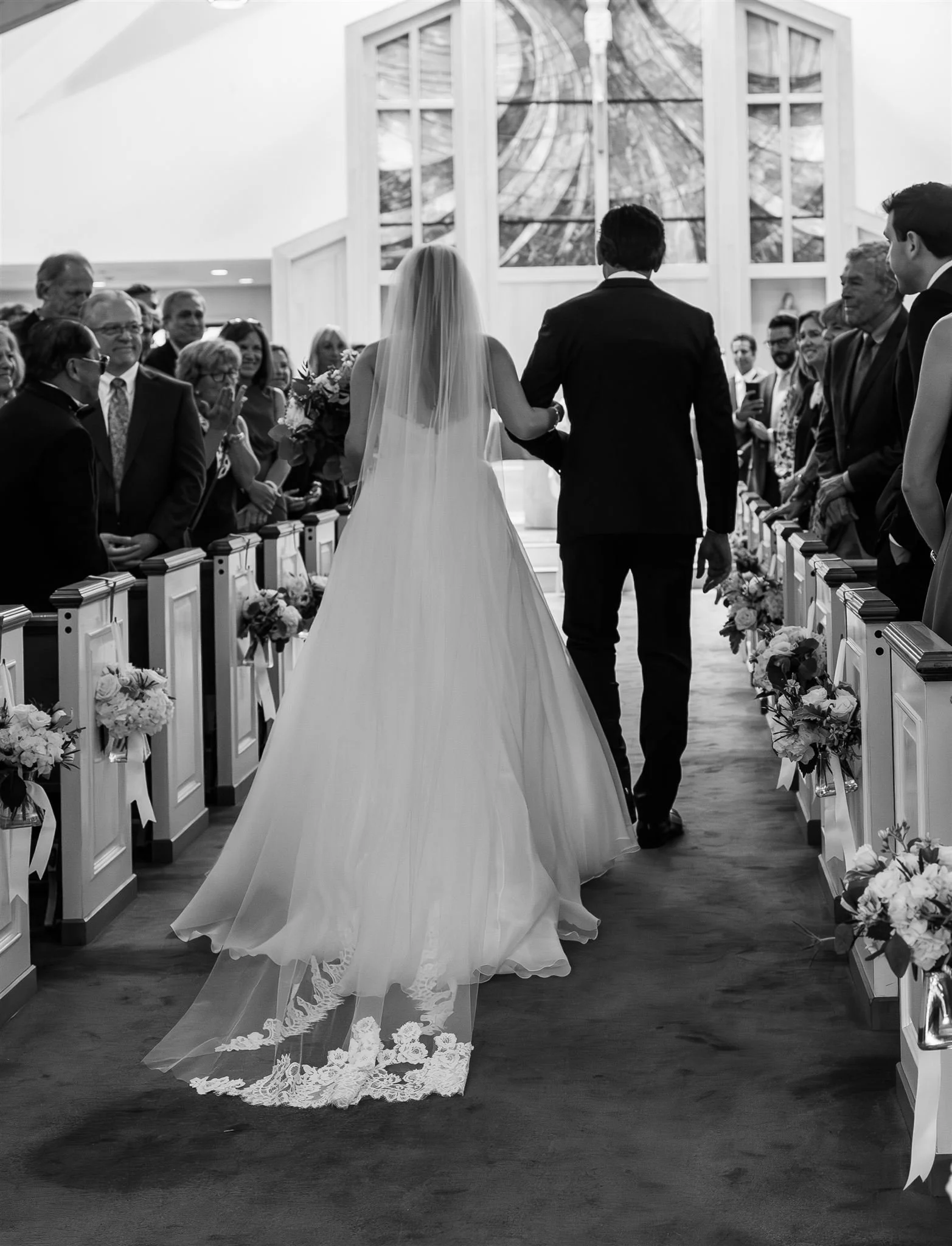 Black and white photograph of a bride and her father walking down the aisle in a church, with wedding guests standing on either side, wearing formal attire, and floral decorations on the pews.