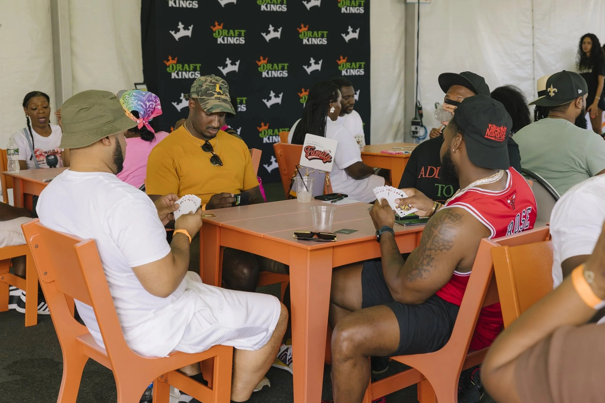 People playing cards at un outdoor event with a black backdrop displaying the Coop Draft Kings logo. The people are seated around orange tables and chairs, engaging in a card game, with some wearing casual attire, hats, and sunglasses.