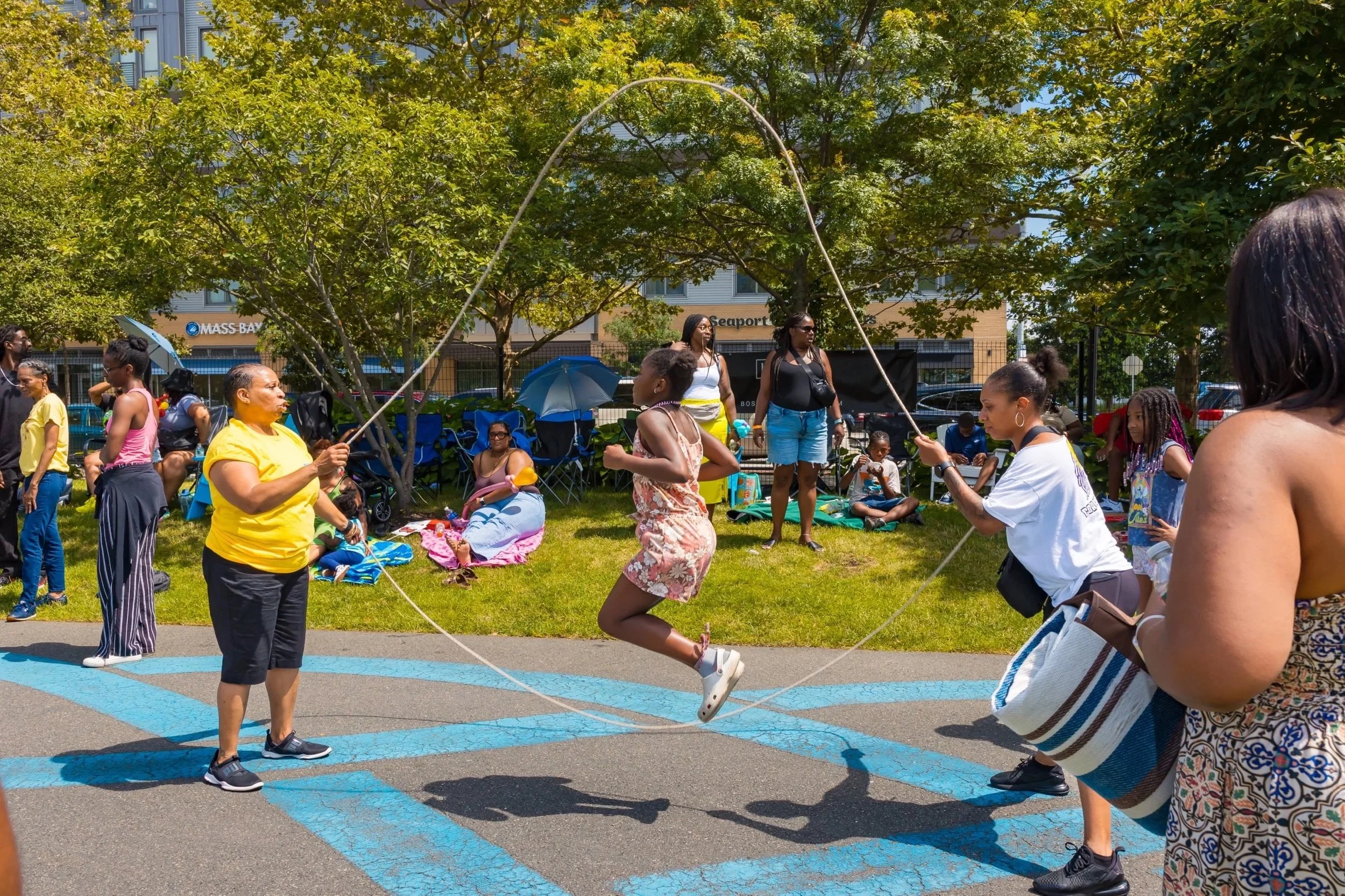 People enjoying a sunny day outdoors, with some sitting on blankets and others watching children jump rope in a park area surrounded by trees and buildings.