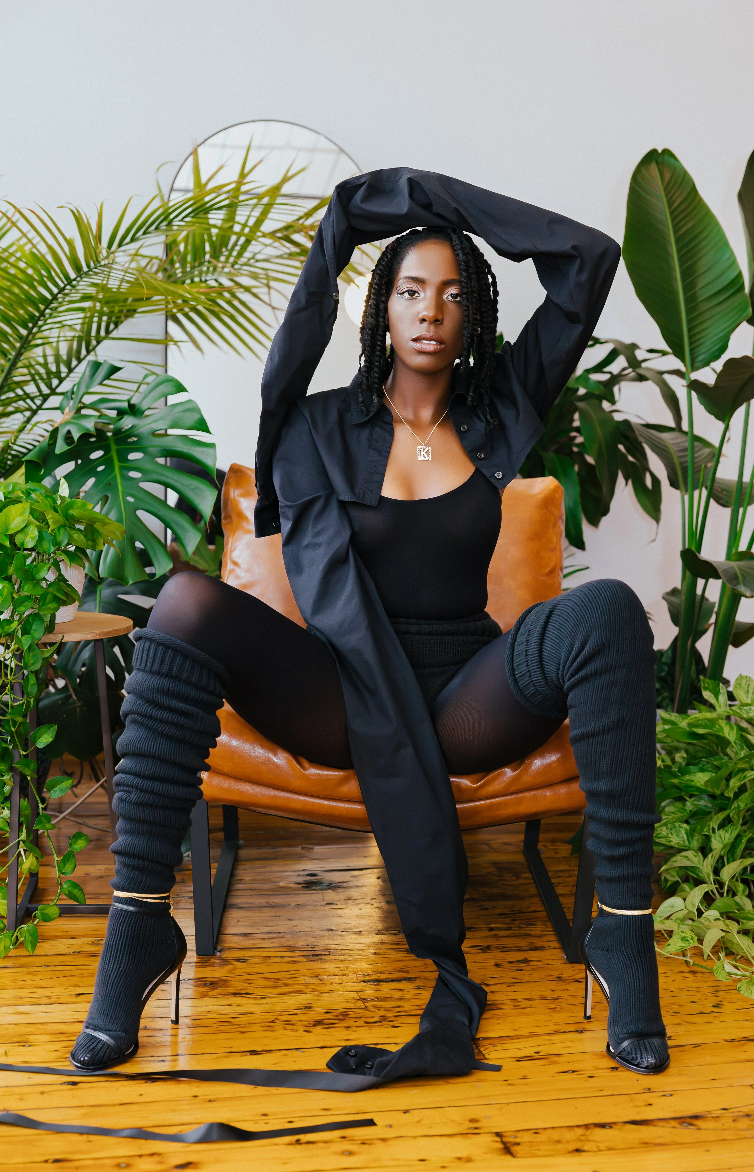 A woman sitting on a brown leather chair surrounded by green indoor plants, wearing a black tank top, black shorts, black thigh-high socks, black high heels, a black shirt, and jewelry, with her hands raised above her head.