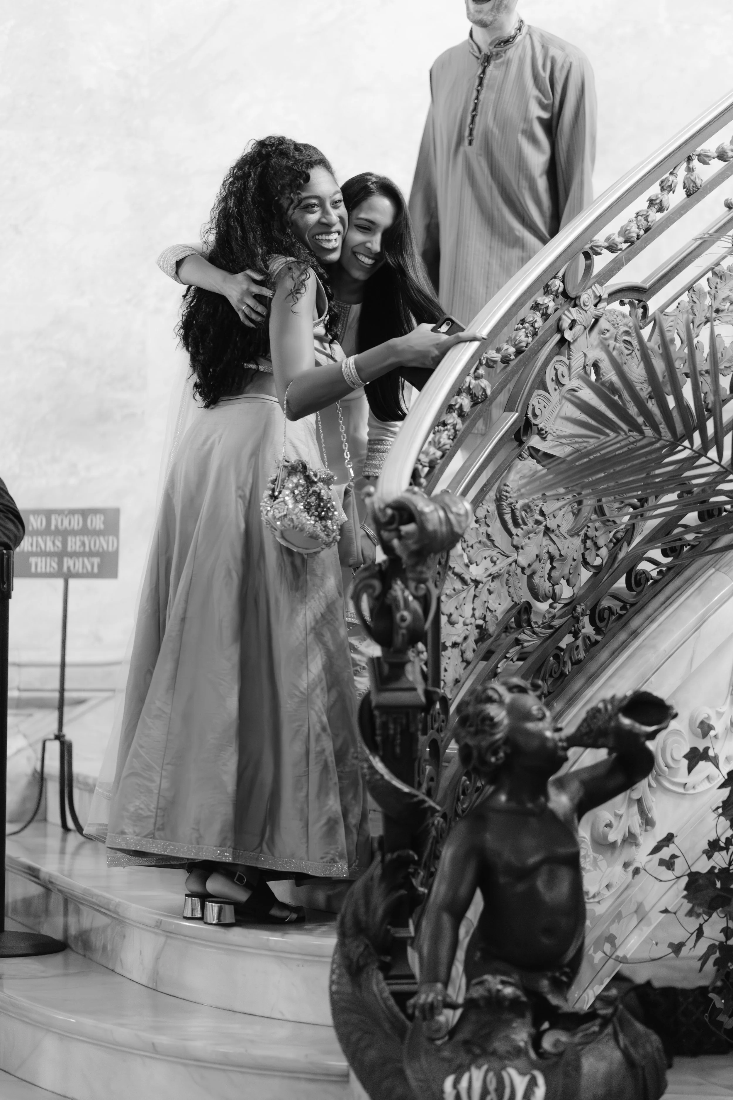 Two women in traditional South Asian attire taking a selfie on an ornate staircase, with one woman hugging the other, and a man standing nearby, in a black-and-white photo.